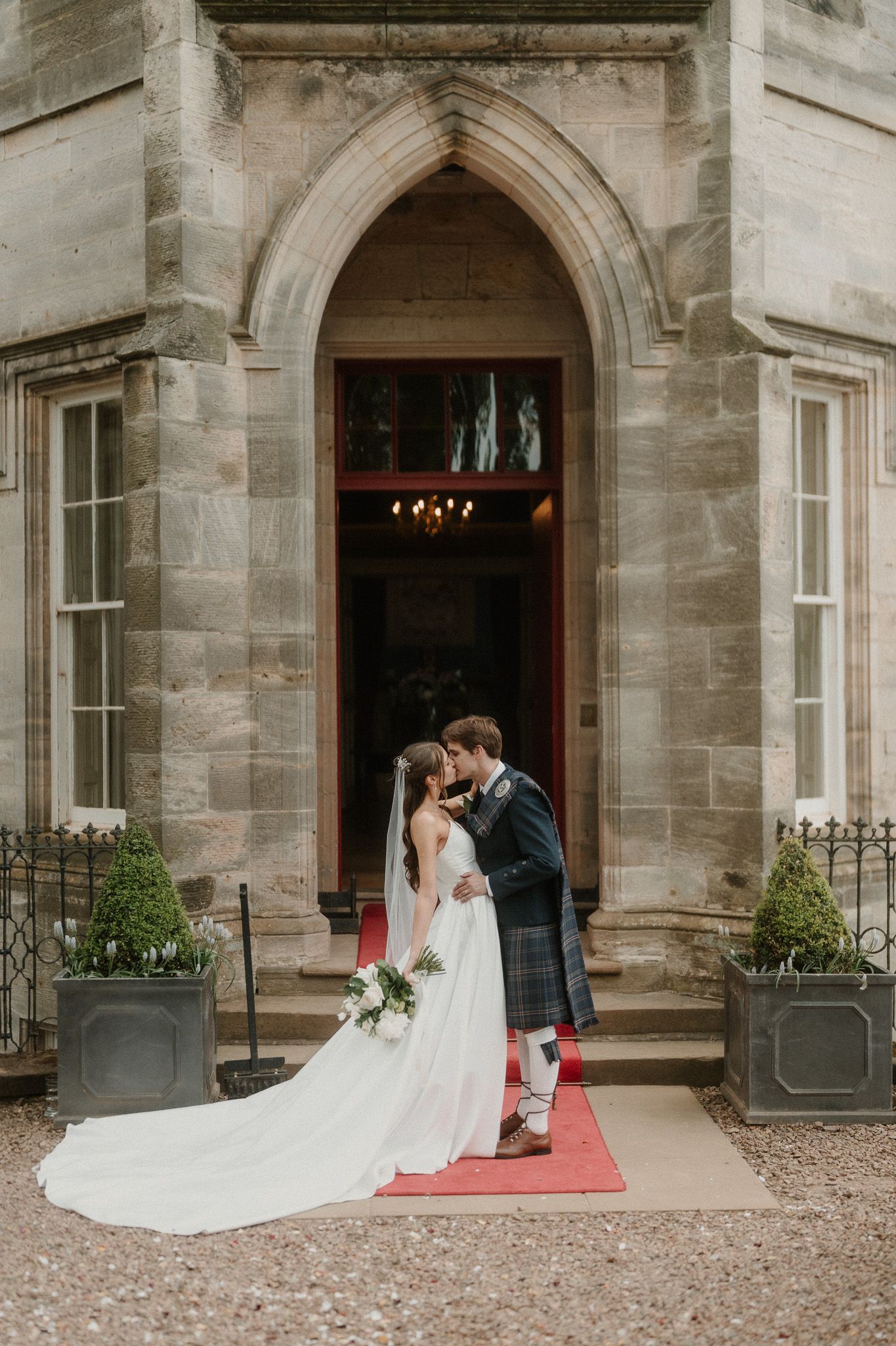 Bride and groom kissing at the entrance of a Scottish stone manor, groom wearing a traditional kilt, bride in a flowing white gown.