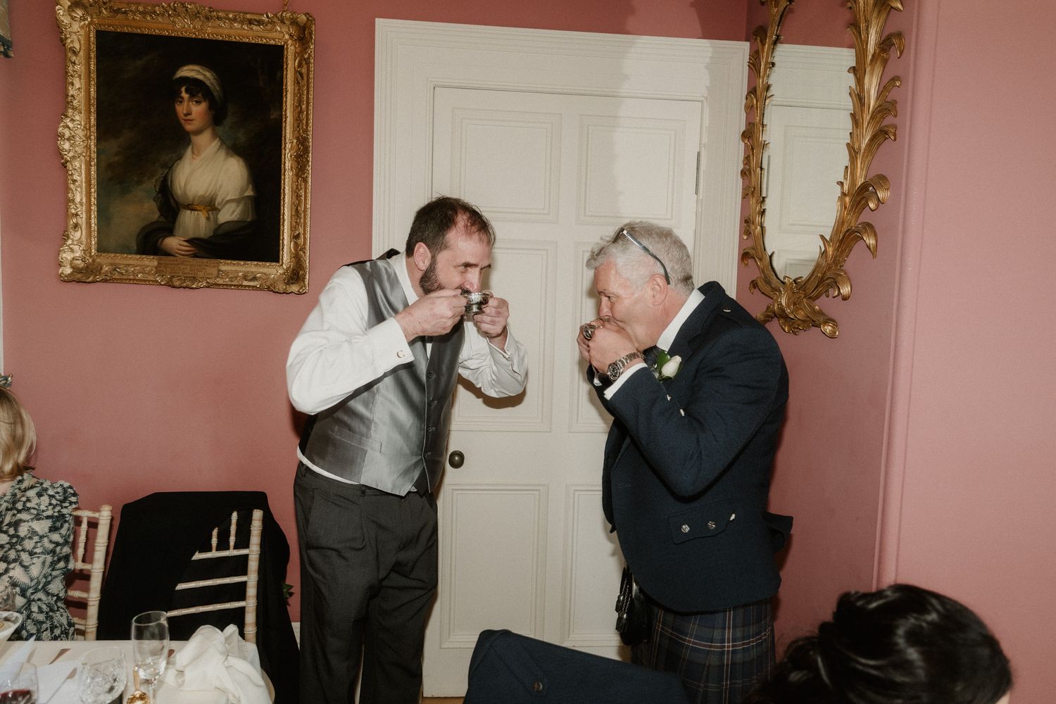 Two men in formal attire share a toast at a wedding reception in an elegant pink-walled room with ornate gold mirrors.