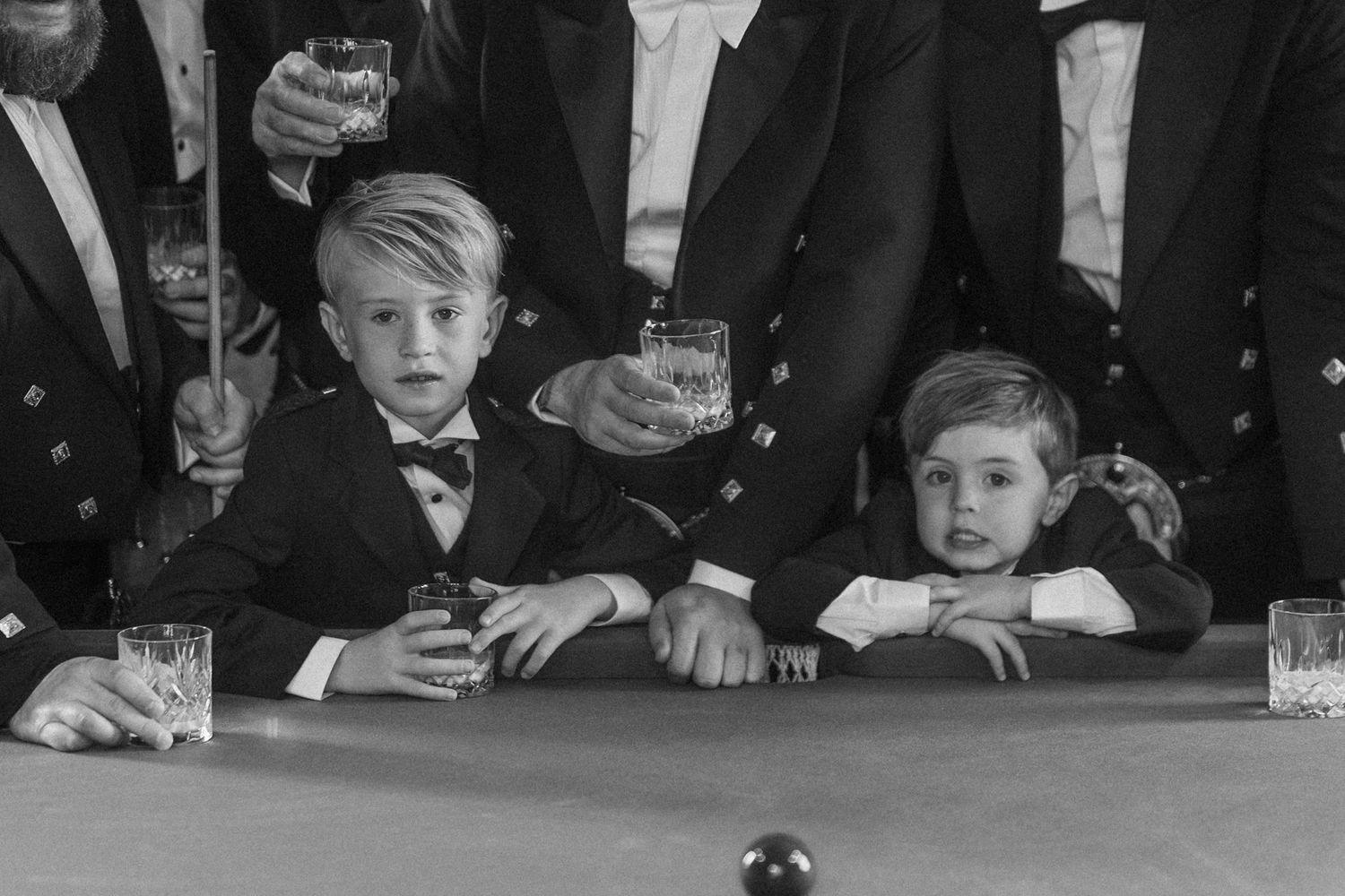Black and white photo of two young boys in suits and bow ties leaning on a pool table as men in formalwear raise their glasses behind them.