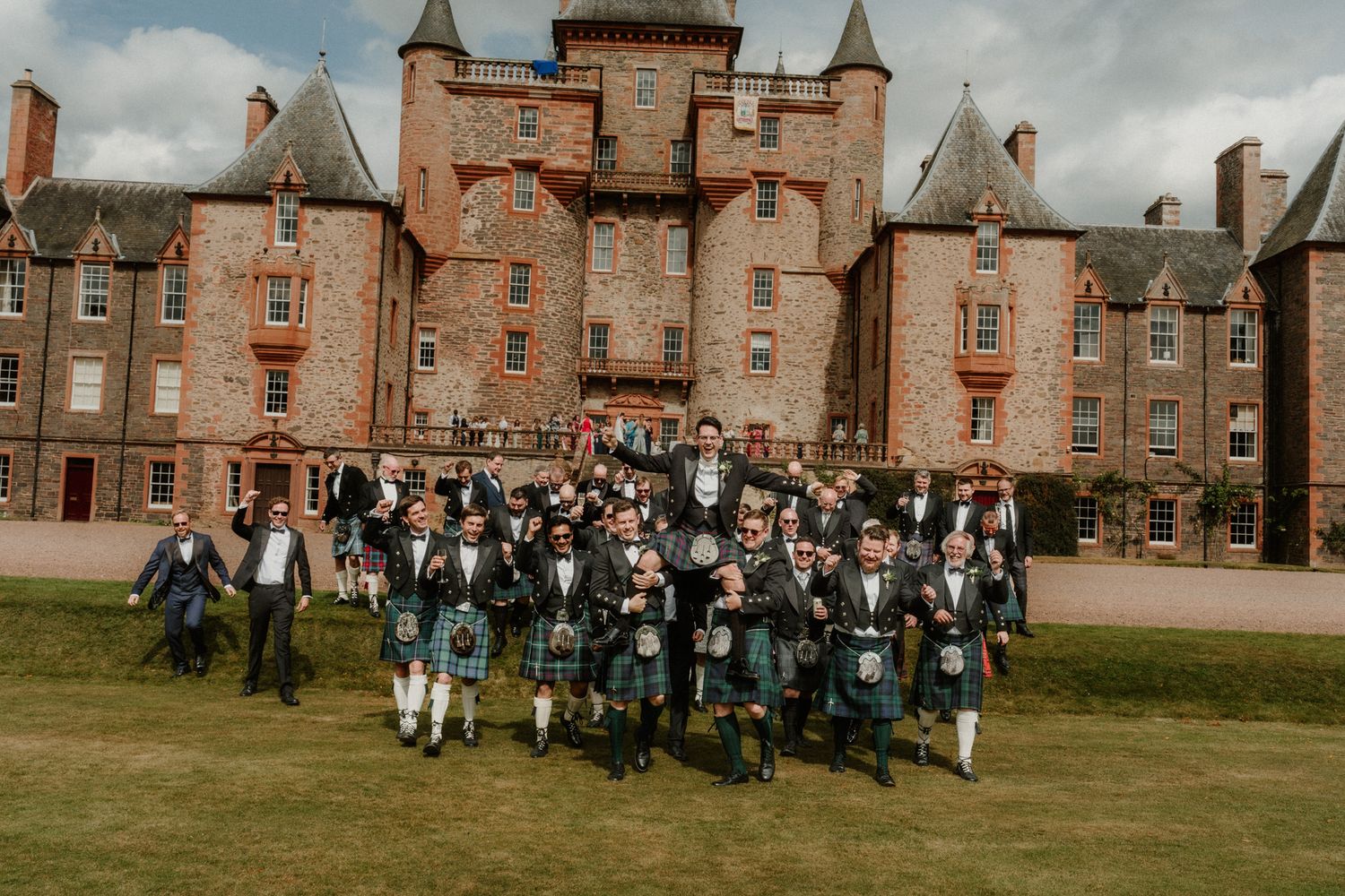Group of men in kilts and formal Highland dress pose on a lawn in front of a historic red sandstone Scottish castle.
