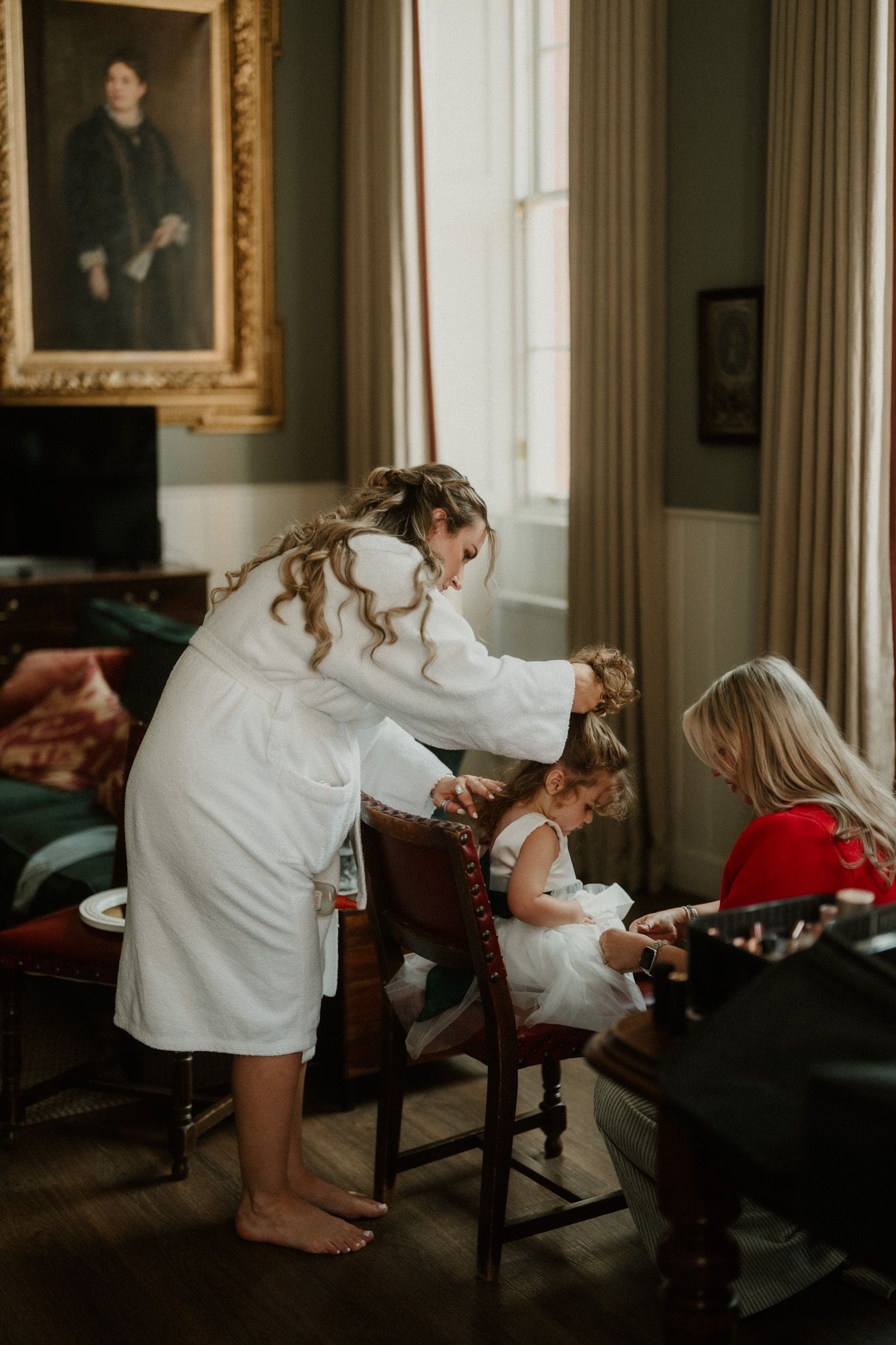 A woman in a white robe leans over to help a flower girl in a white dress get ready beside a seated woman in a elegant, antique-filled room.