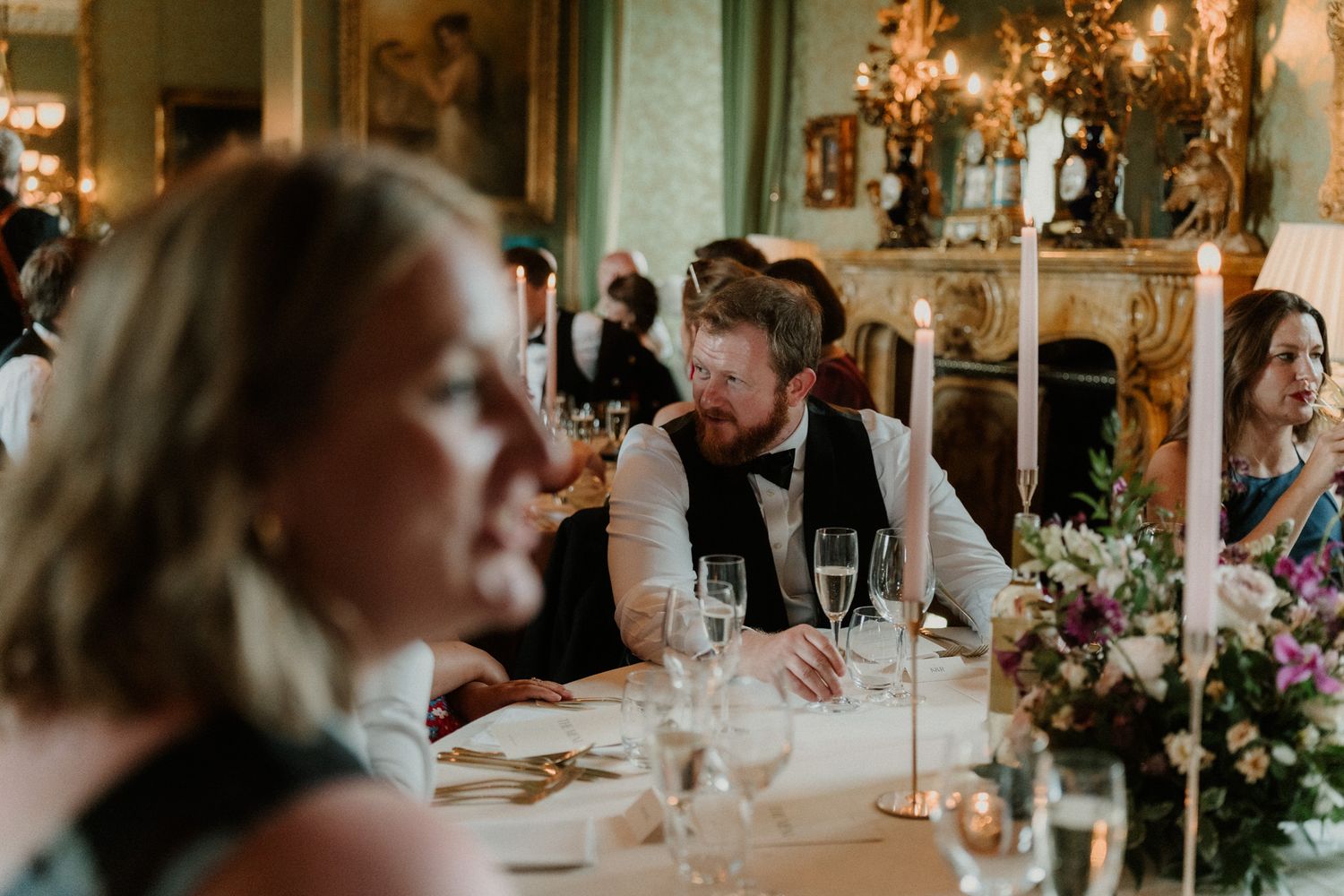 Bearded man in a vest and bow tie smiles at a candlelit wedding reception table with glasses, flowers and guests in an elegant ballroom.