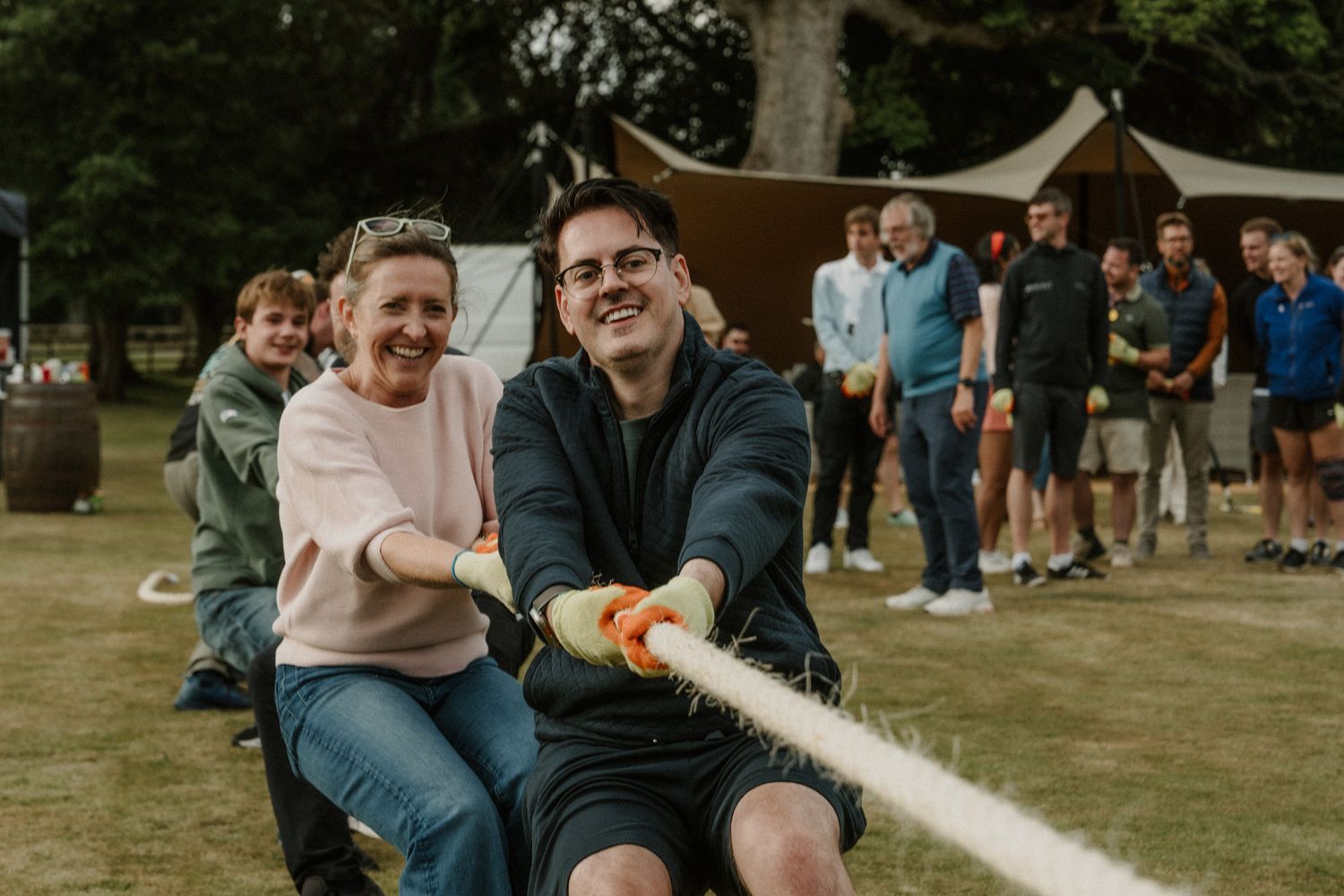 A smiling man and woman pull a thick rope during an outdoor tug of war game while a crowd watches on a grassy field near a tent.