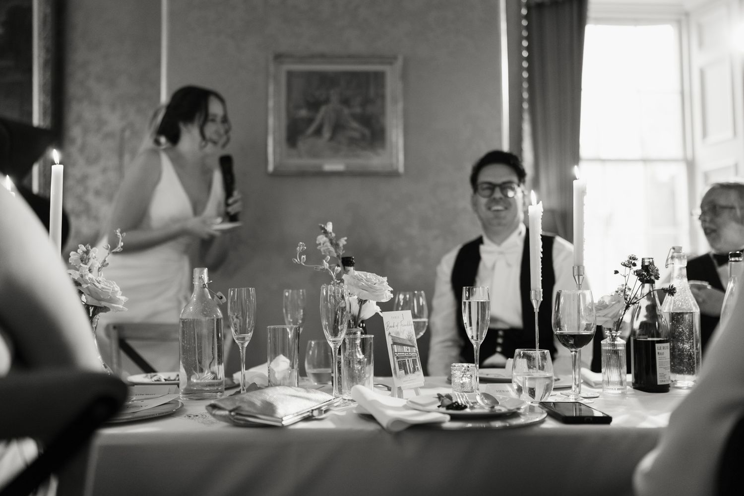 Woman in a white dress speaks with a microphone while smiling guests sit at a candlelit wedding reception table set with glasses.