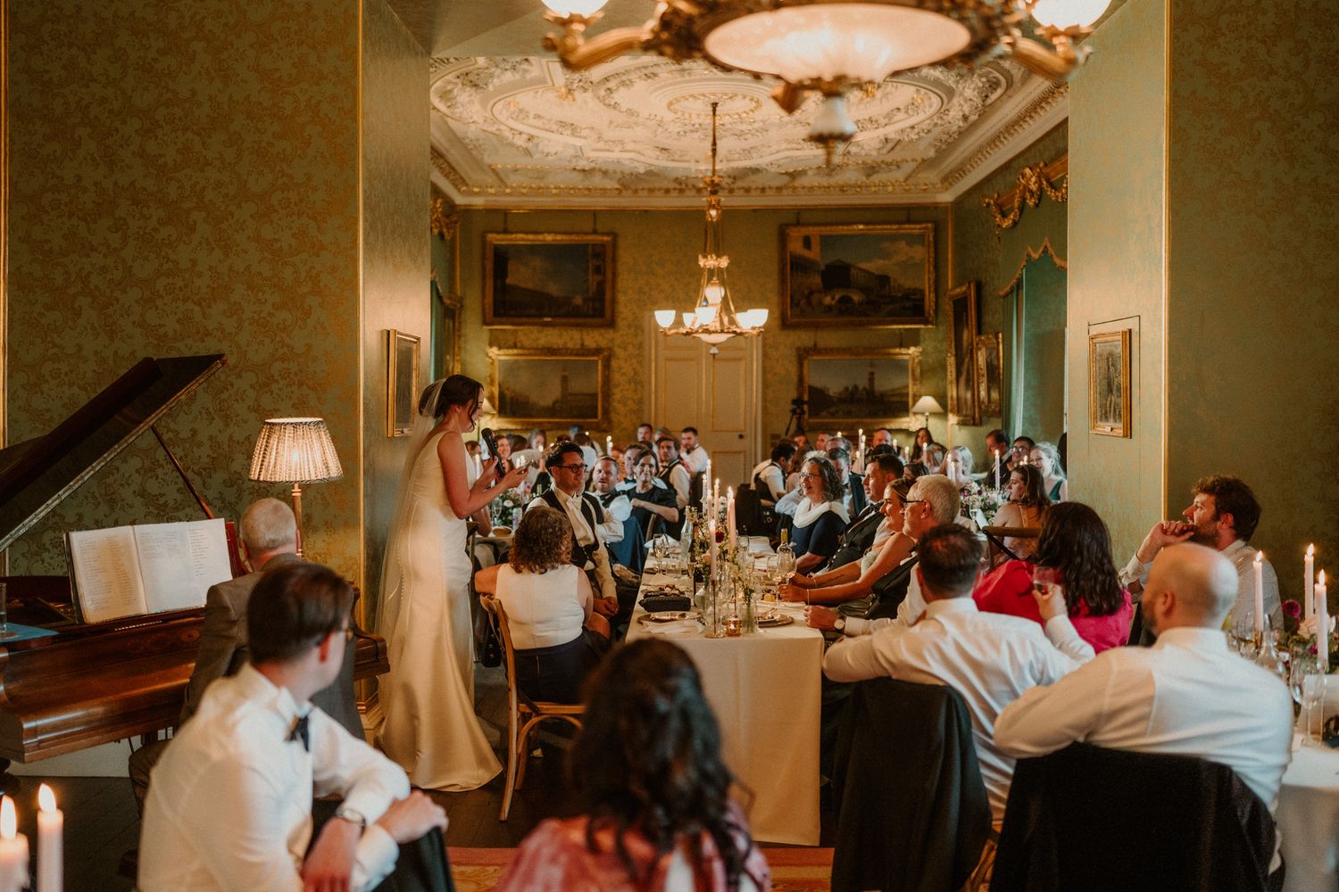 Wedding guests seated at long tables in an elegant dining hall with chandeliers and a grand piano as a woman in white gives a speech.