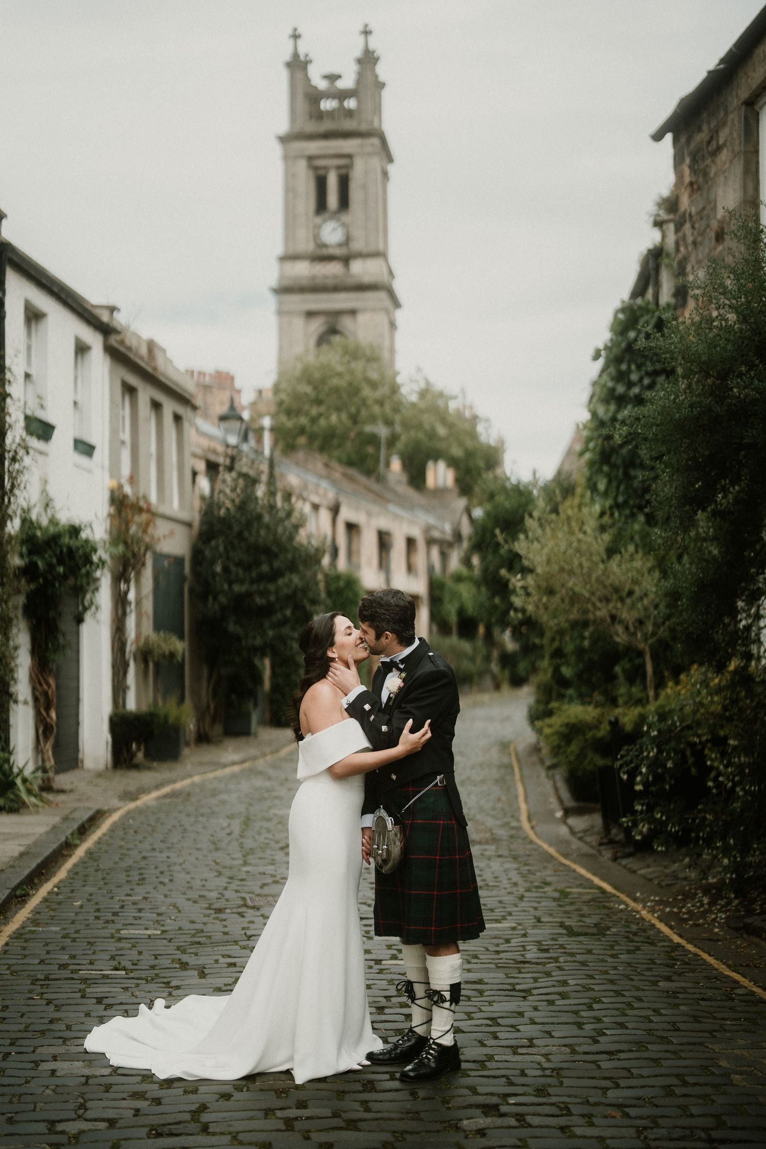 Wedding couple in white gown and traditional kilt share romantic moment on cobblestone lane.