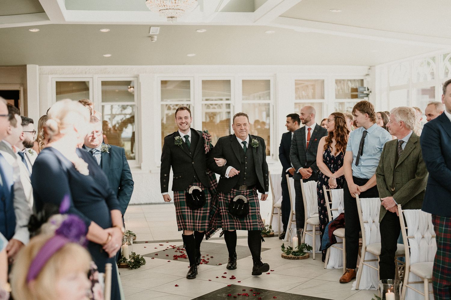Wedding ceremony guests stand in a bright white reception hall.