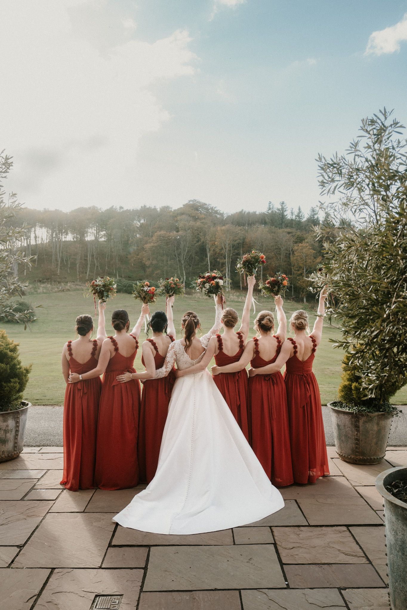 Wedding party in red dresses poses with backs turned showing dress details against scenic backdrop.