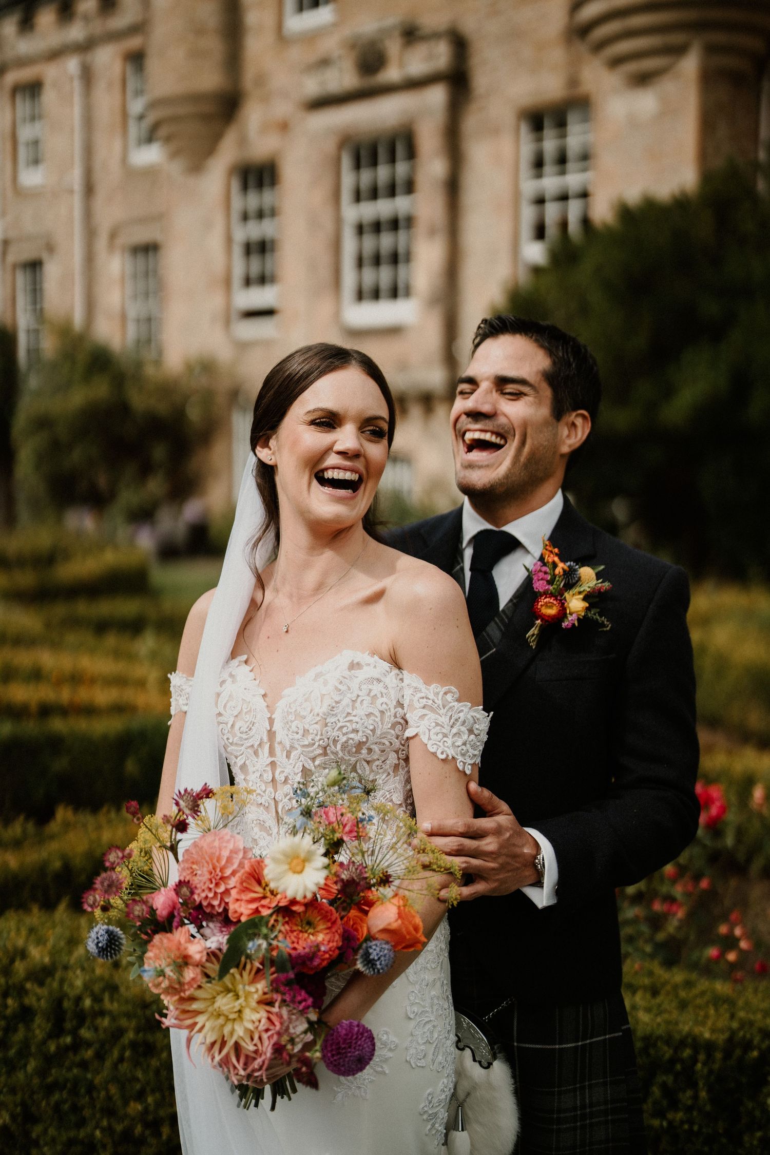 Newlyweds share joyful laughter with vibrant autumn bouquet against stone building.