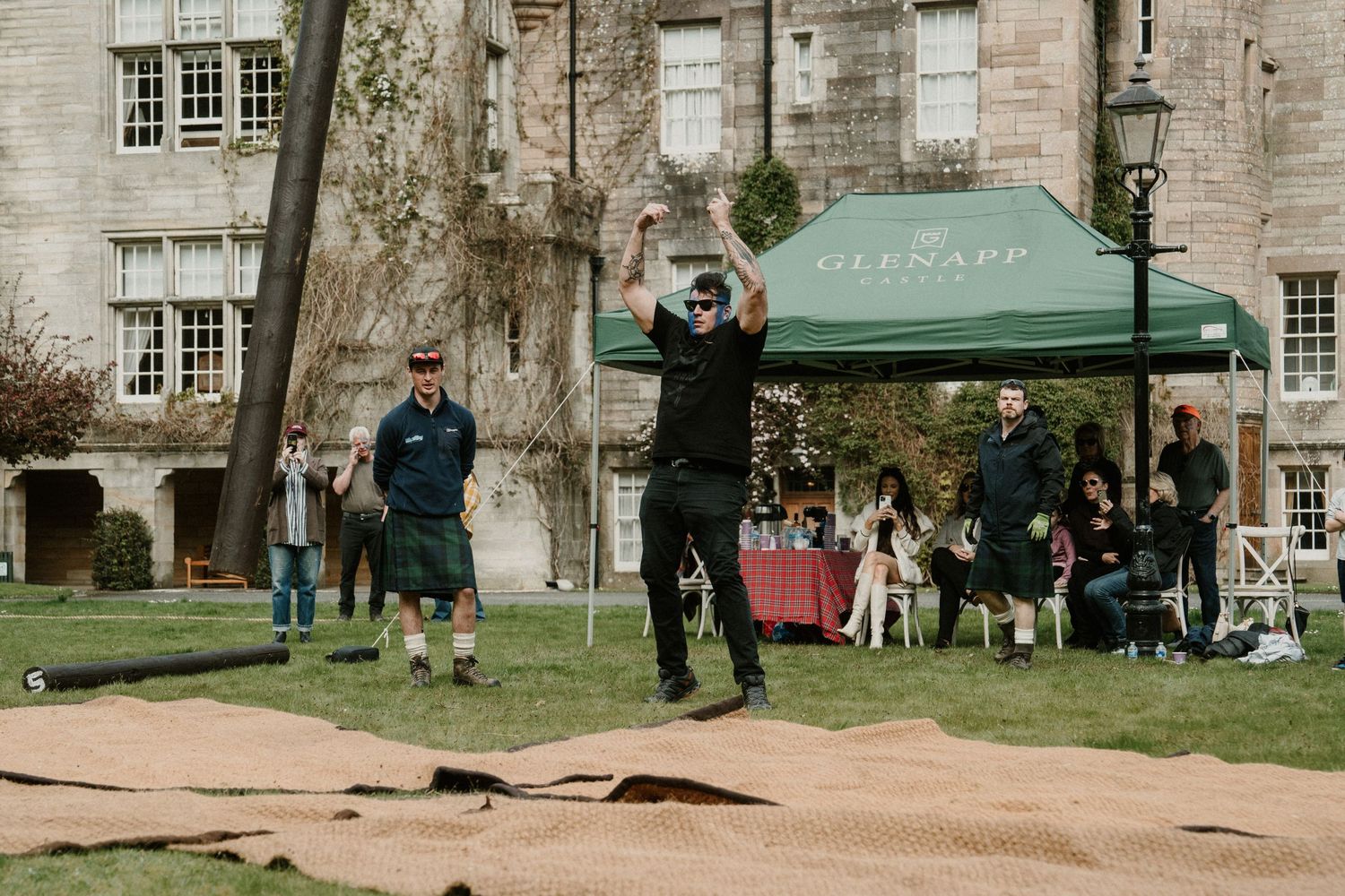 Someone demonstrates axe throwing technique under a green tent near a stone building.