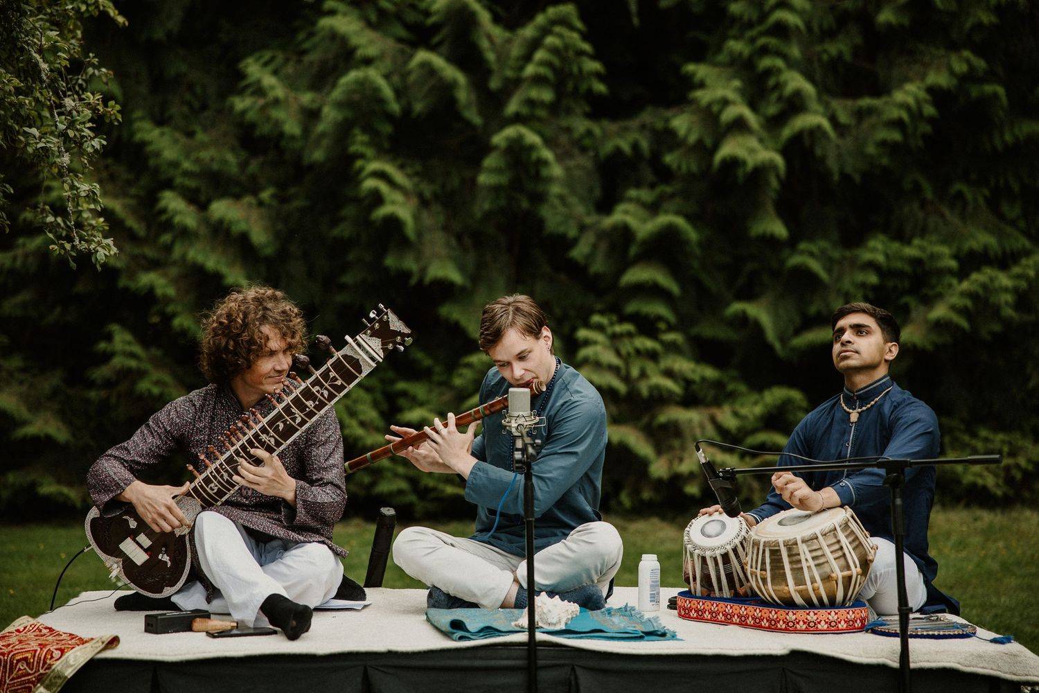 Musicians playing traditional Indian instruments while seated outdoors among greenery.