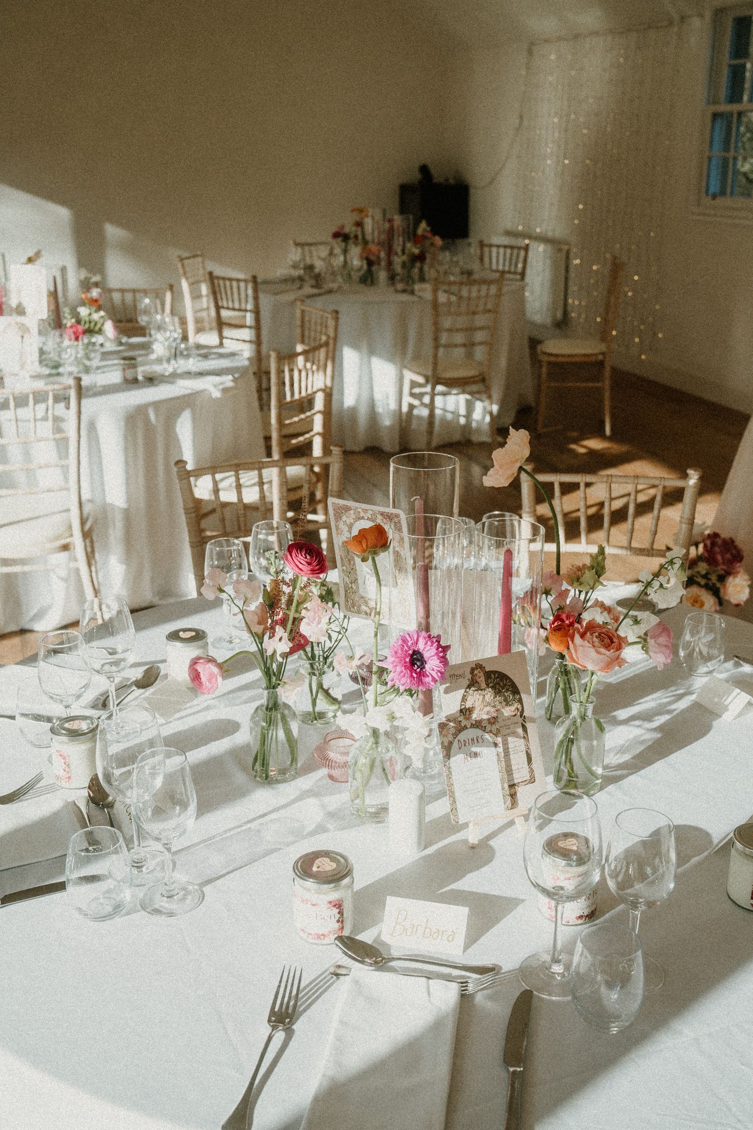 Elegant wedding reception table setting with pink floral centerpieces and white linens in natural sunlight.