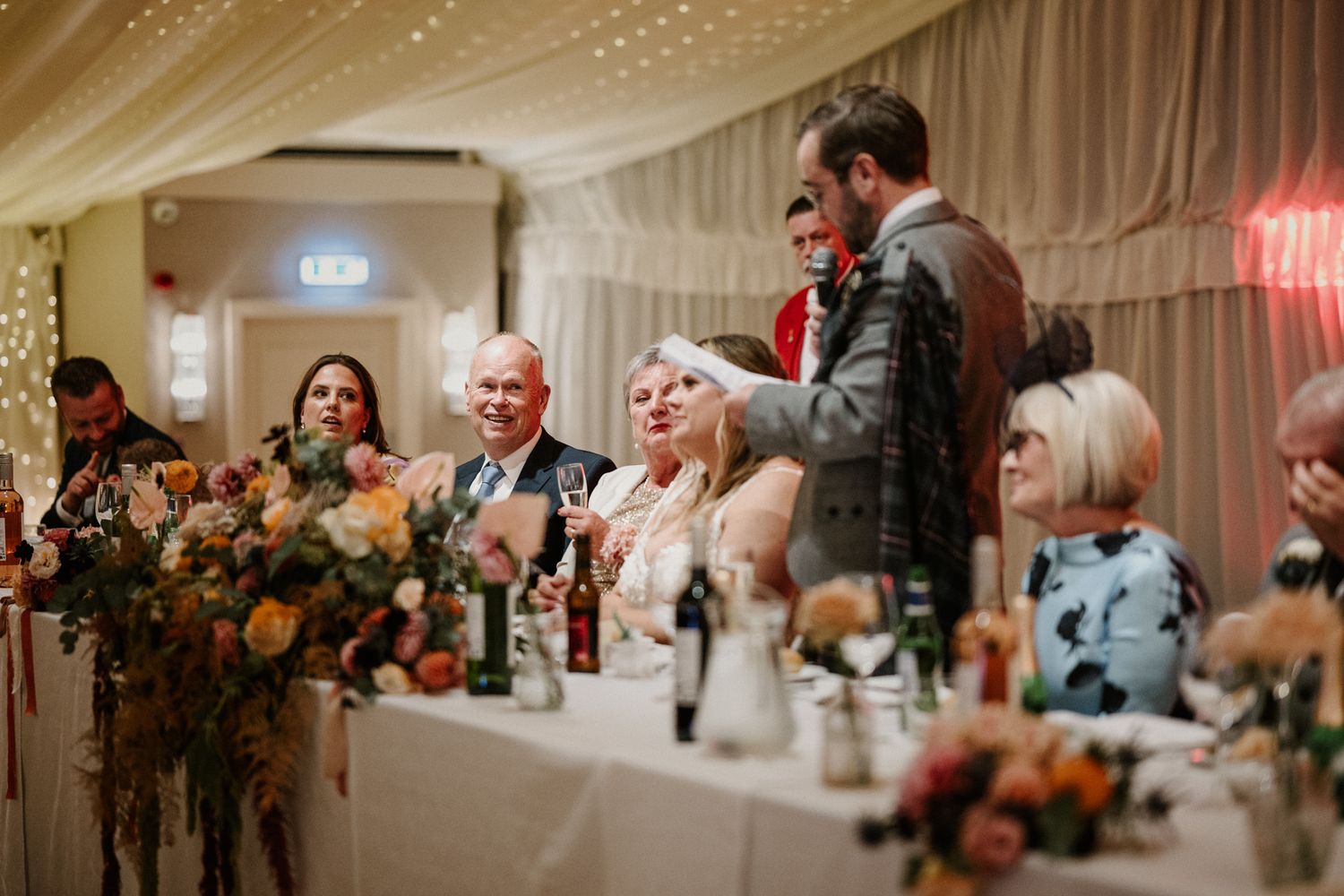 Wedding guests enjoying speeches at a reception with elegant floral arrangements and draped decor.
