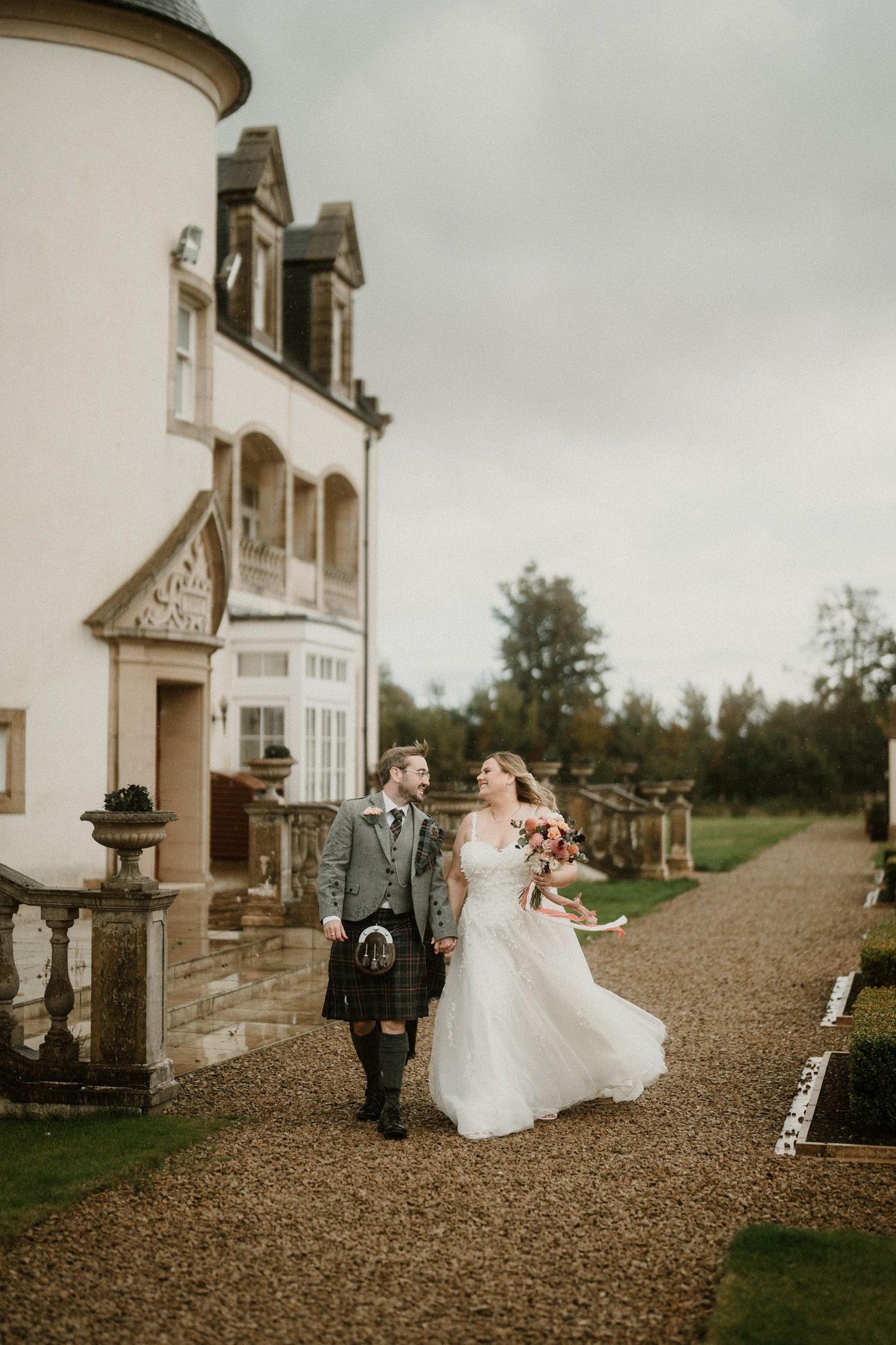 A wedding couple poses outside a grand white building on a gravel path.