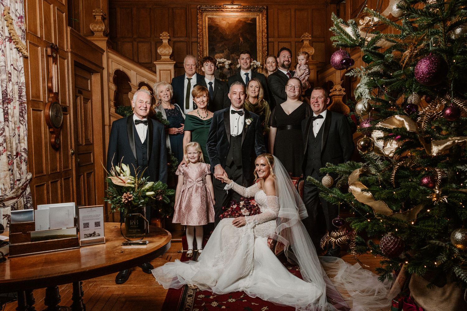 Formal wedding party portrait on historic staircase with decorated Christmas tree and wood paneling.