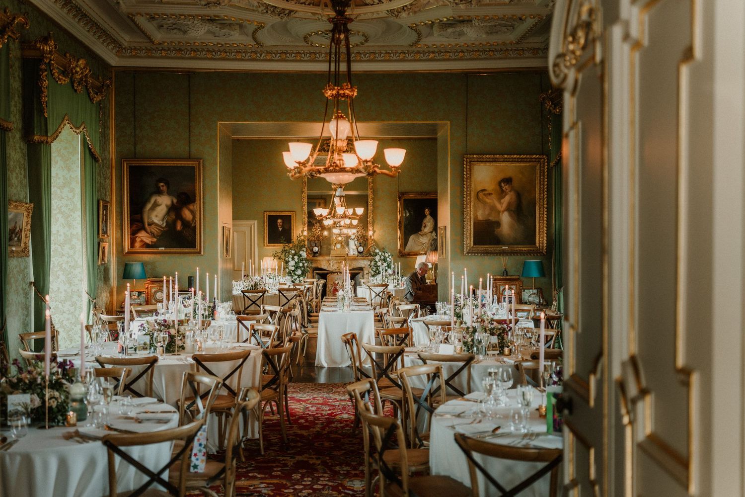 Elegant historic dining room with set tables, ornate chandeliers, framed oil paintings, green walls and a rich patterned carpet.