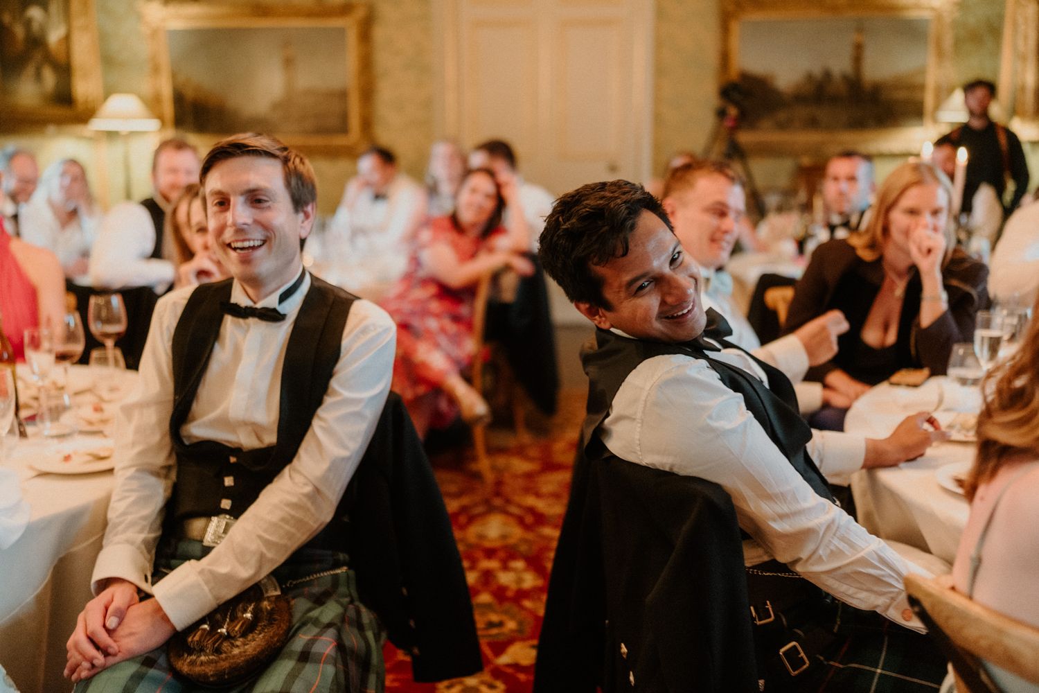 Two smiling men in kilts and waistcoats laugh while seated at a formal dinner with guests at tables in an elegant, ornate hall.
