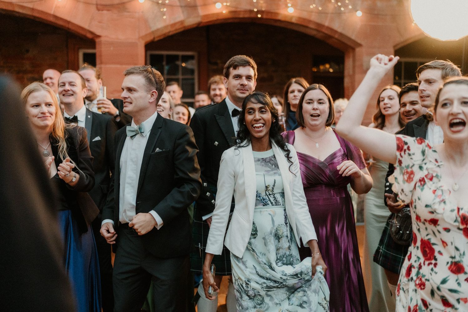 Joyful wedding guests in formal attire laugh and cheer together at an evening celebration in front of a warmly lit brick building.