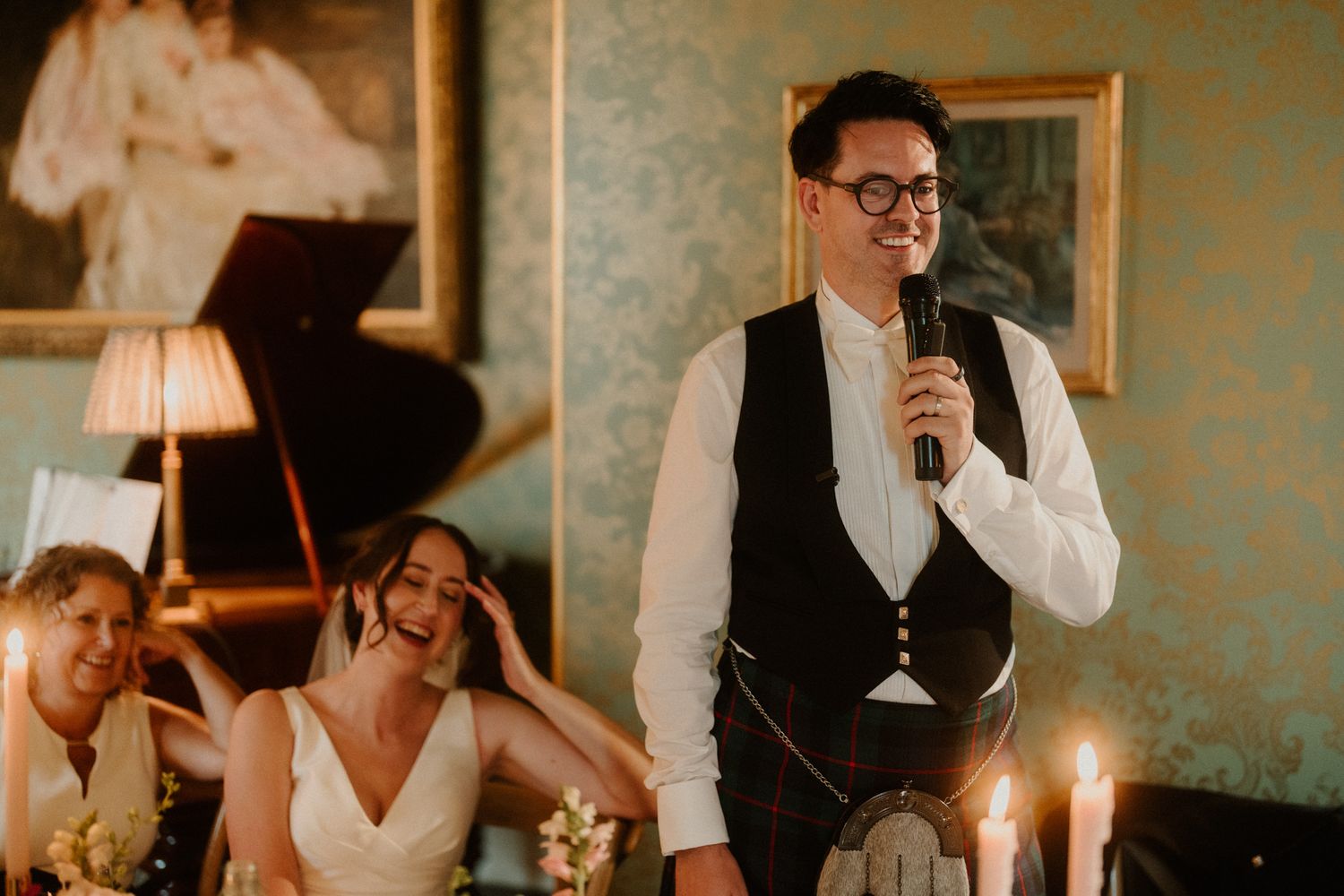 Man in glasses, vest and kilt gives a wedding speech with a microphone as a smiling bride laughs beside candlelit tables.
