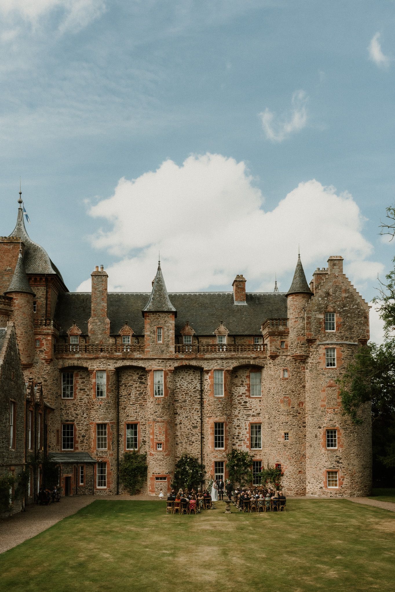 Wedding guests gather on a manicured lawn for an outdoor ceremony in front of a grand stone castle with turrets under a blue sky.
