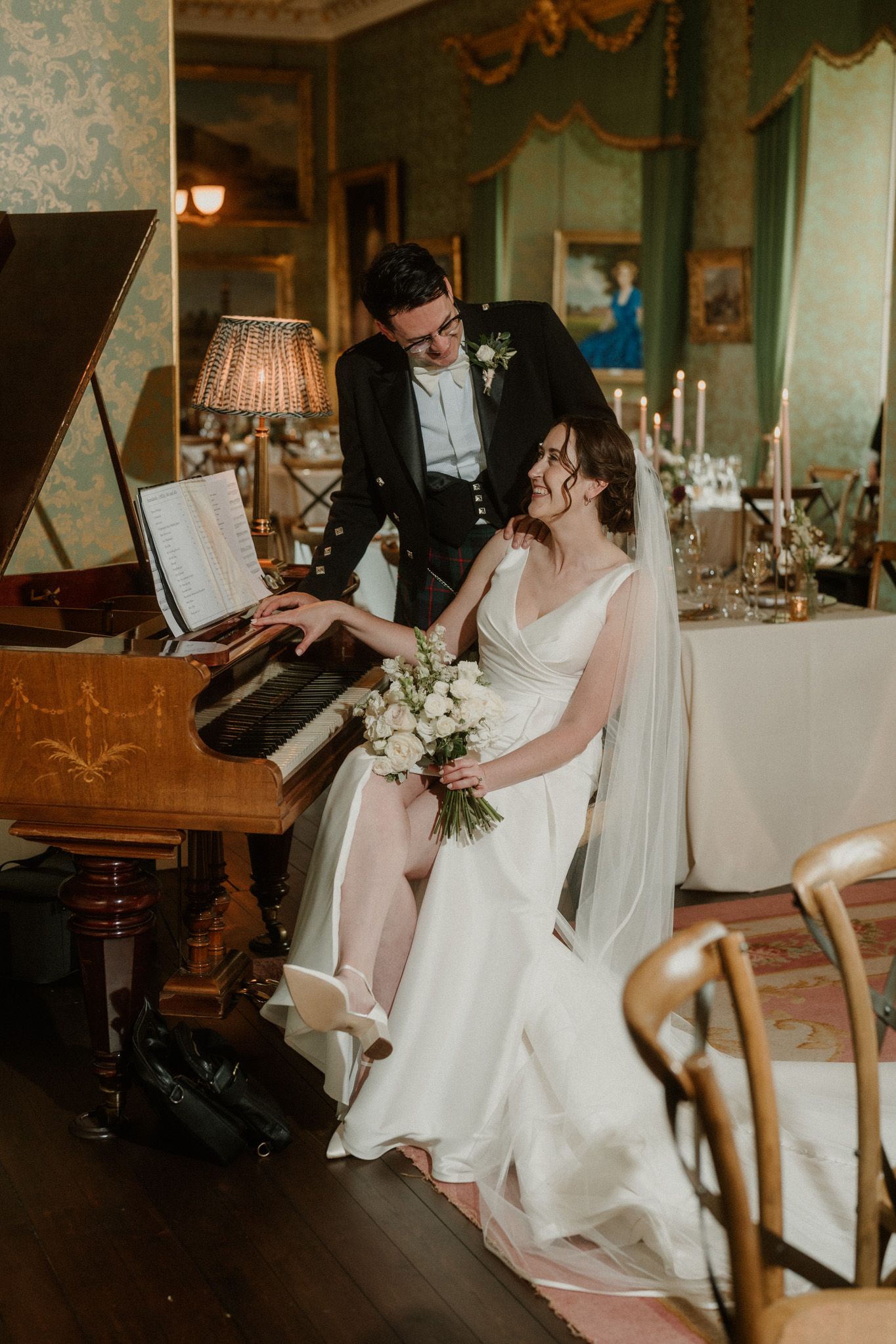 Bride in a white gown with veil and bouquet sits at a vintage piano beside her groom in an elegant candlelit wedding venue.