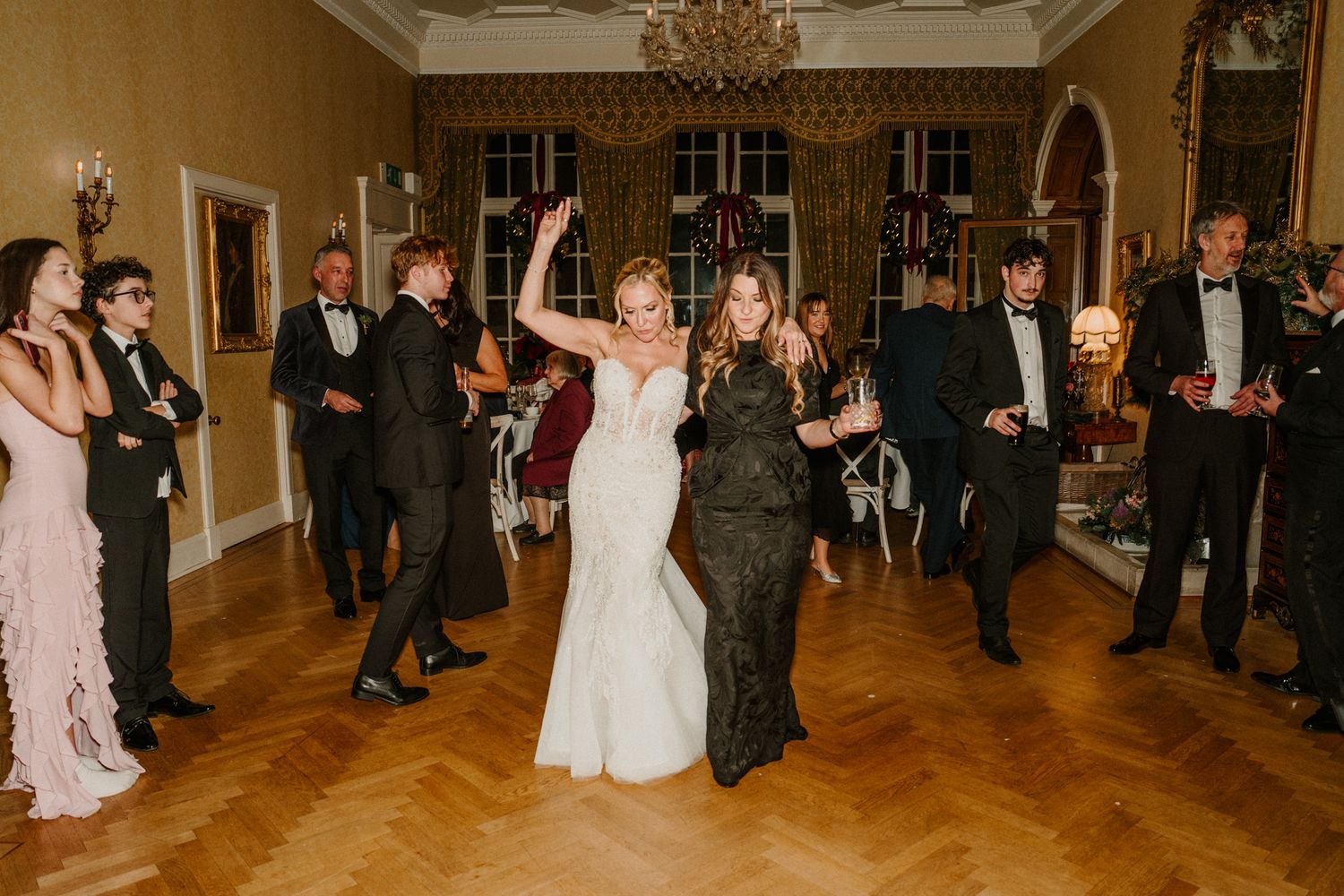 Wedding guests dance and celebrate on a polished wooden floor beneath crystal chandeliers.