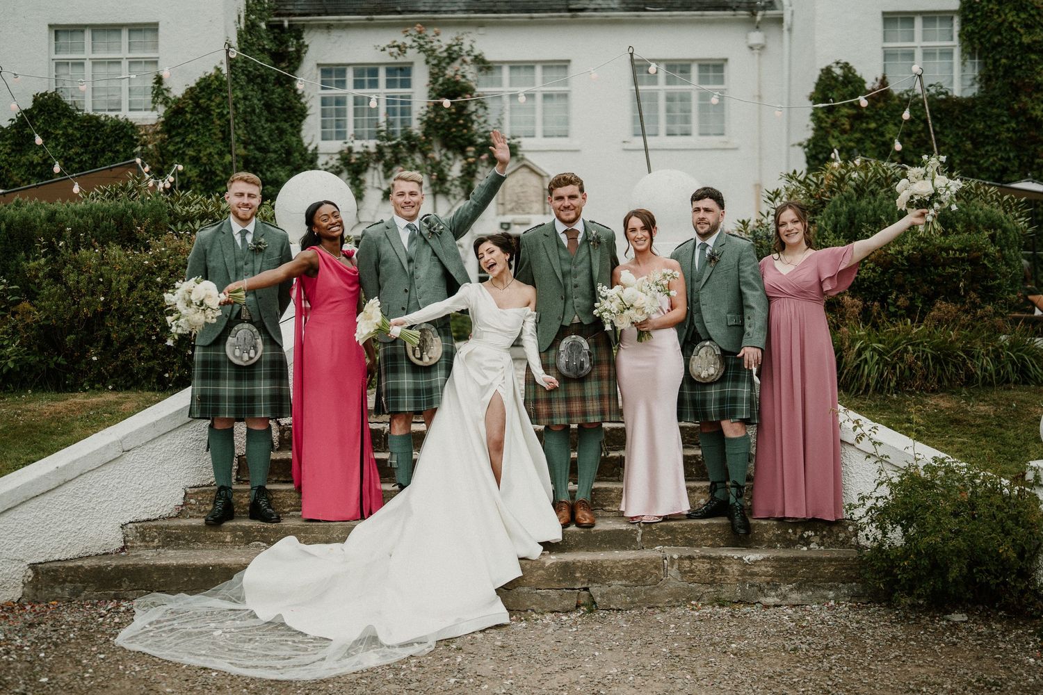 Wedding party group photo with bridesmaids in pink dresses and groomsmen in traditional Scottish attire.