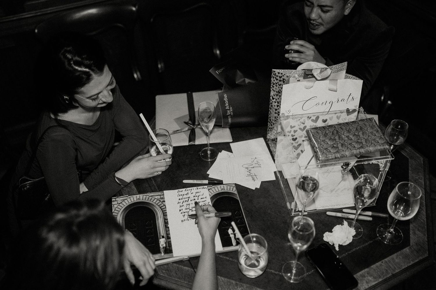 Dark moody reception scene with guests at tables writing and socializing.