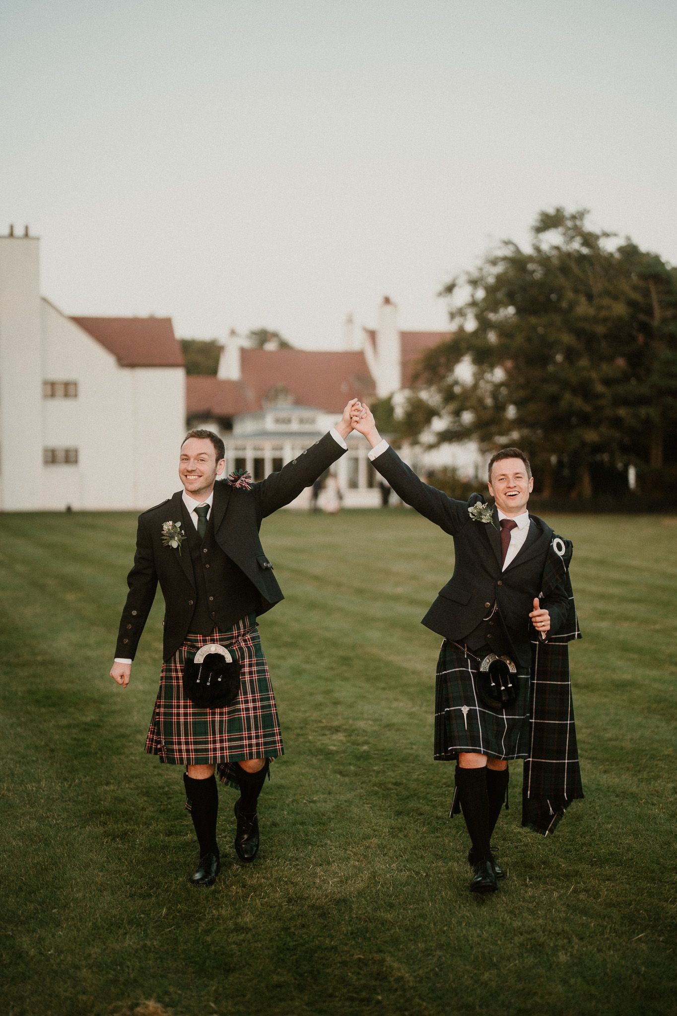 Two people in traditional Scottish kilts celebrating on a grassy field with buildings in background.
