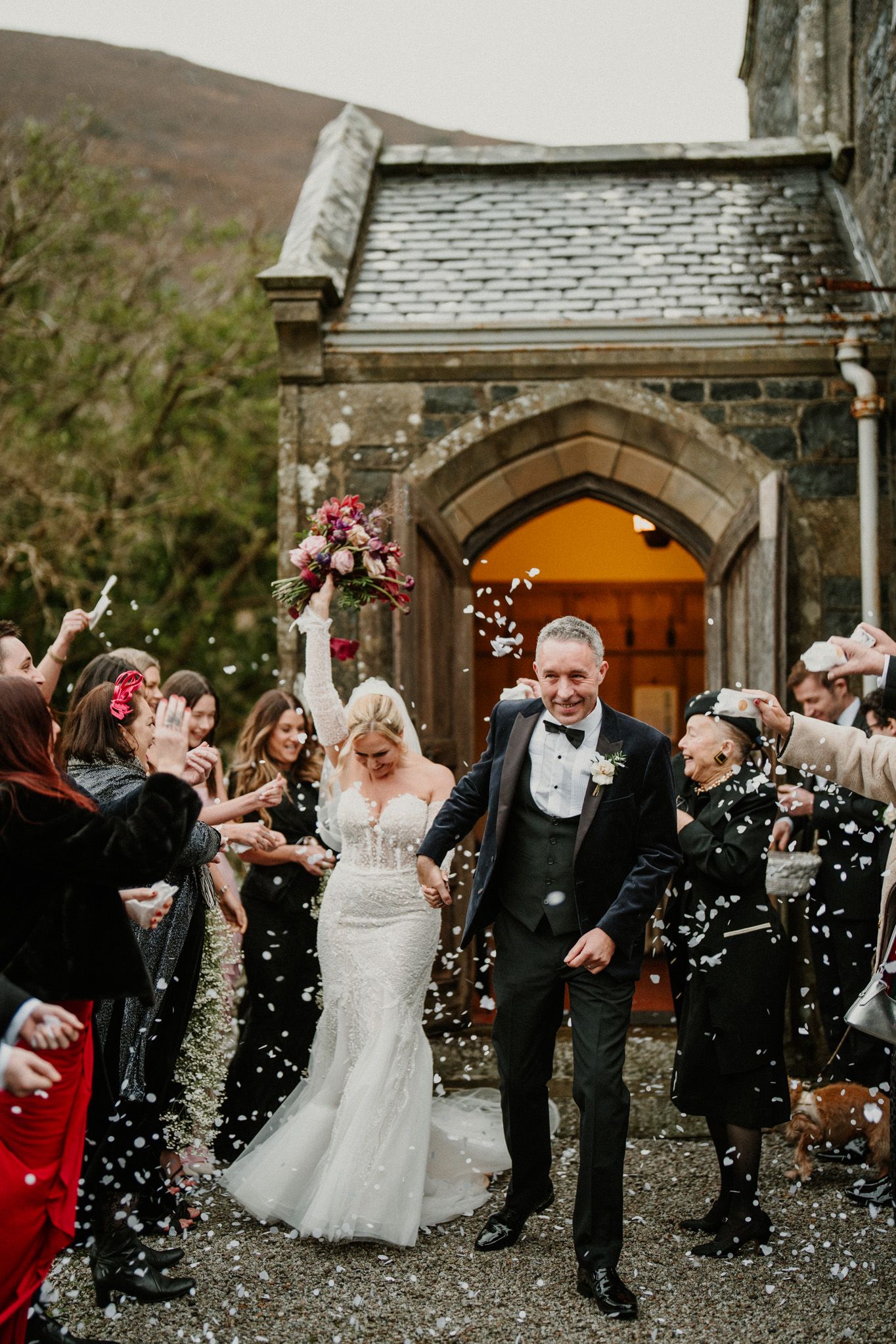 Joyful wedding exit with guests throwing confetti outside stone church entrance.