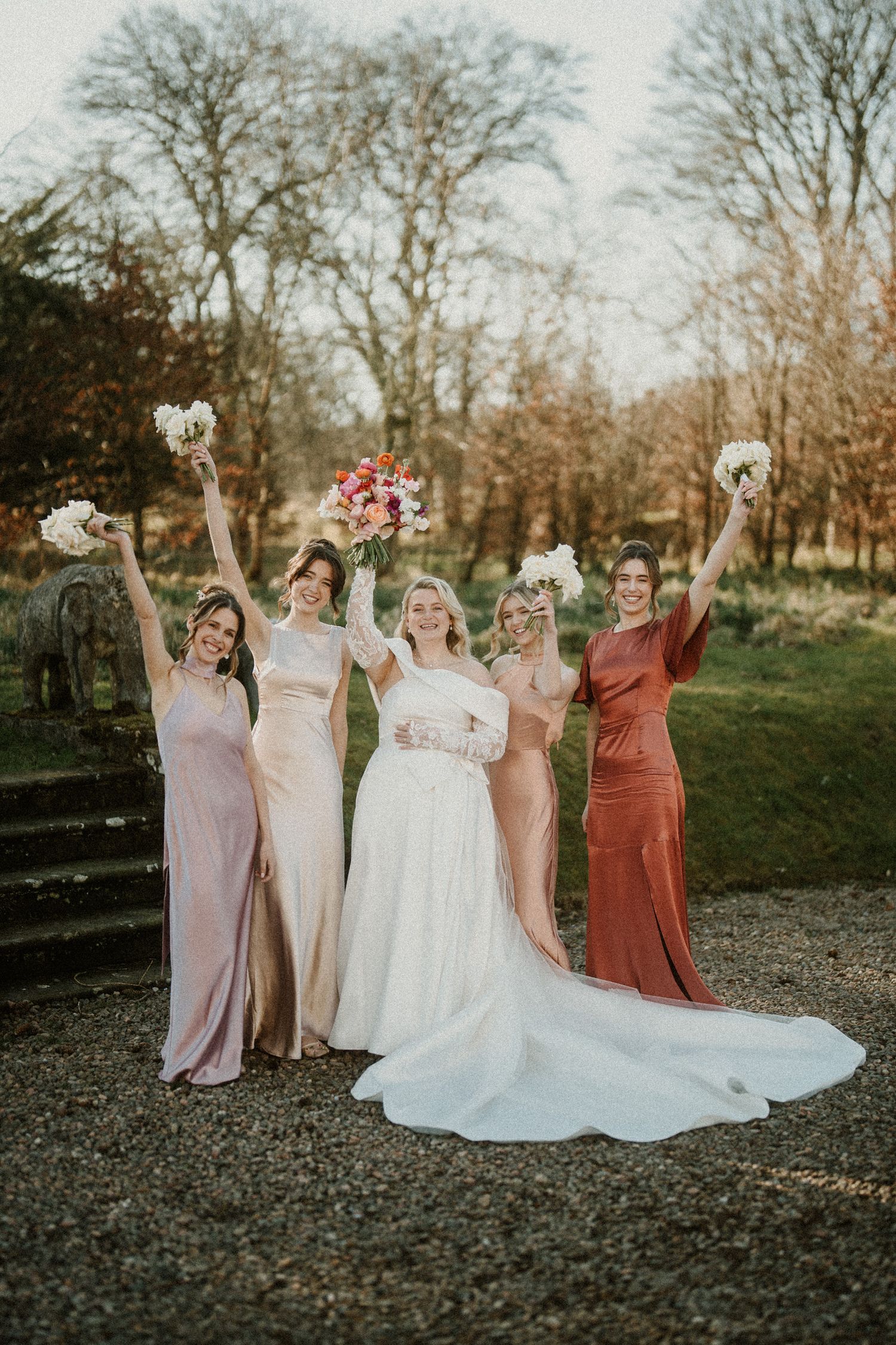 Bridesmaids in dusty rose and terracotta dresses gather around bride for outdoor wedding photos.