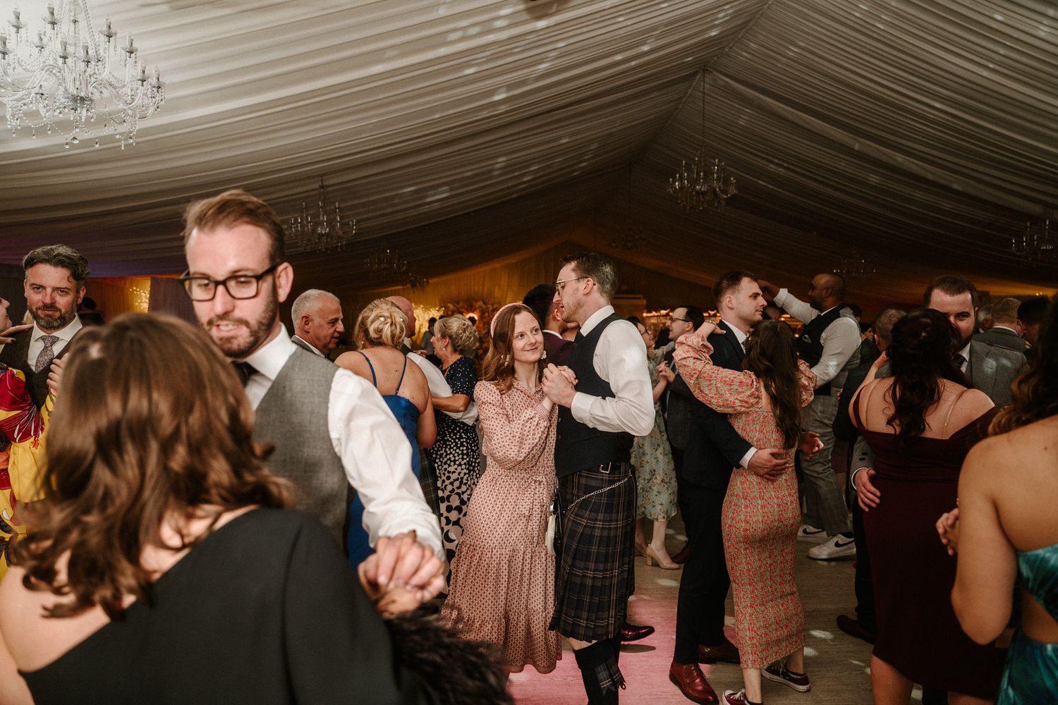 Wedding guests dancing and celebrating on a lively dance floor under draped white ceiling.
