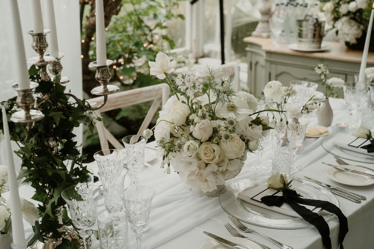 Elegant wedding table setting with white flowers and greenery in a greenhouse venue.