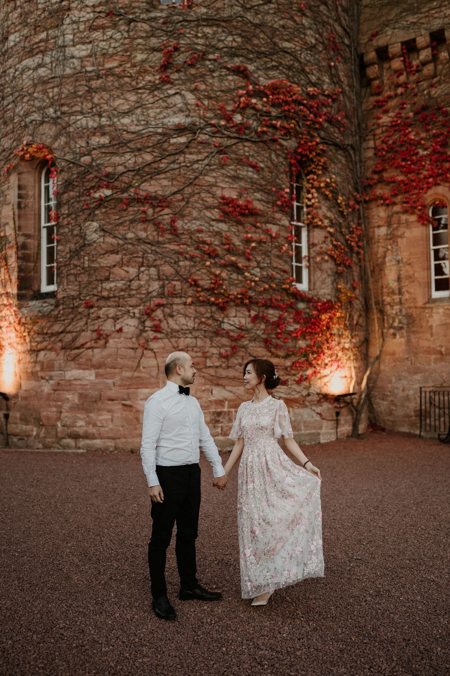 A couple walks hand in hand along a stone wall covered in red autumn vines.