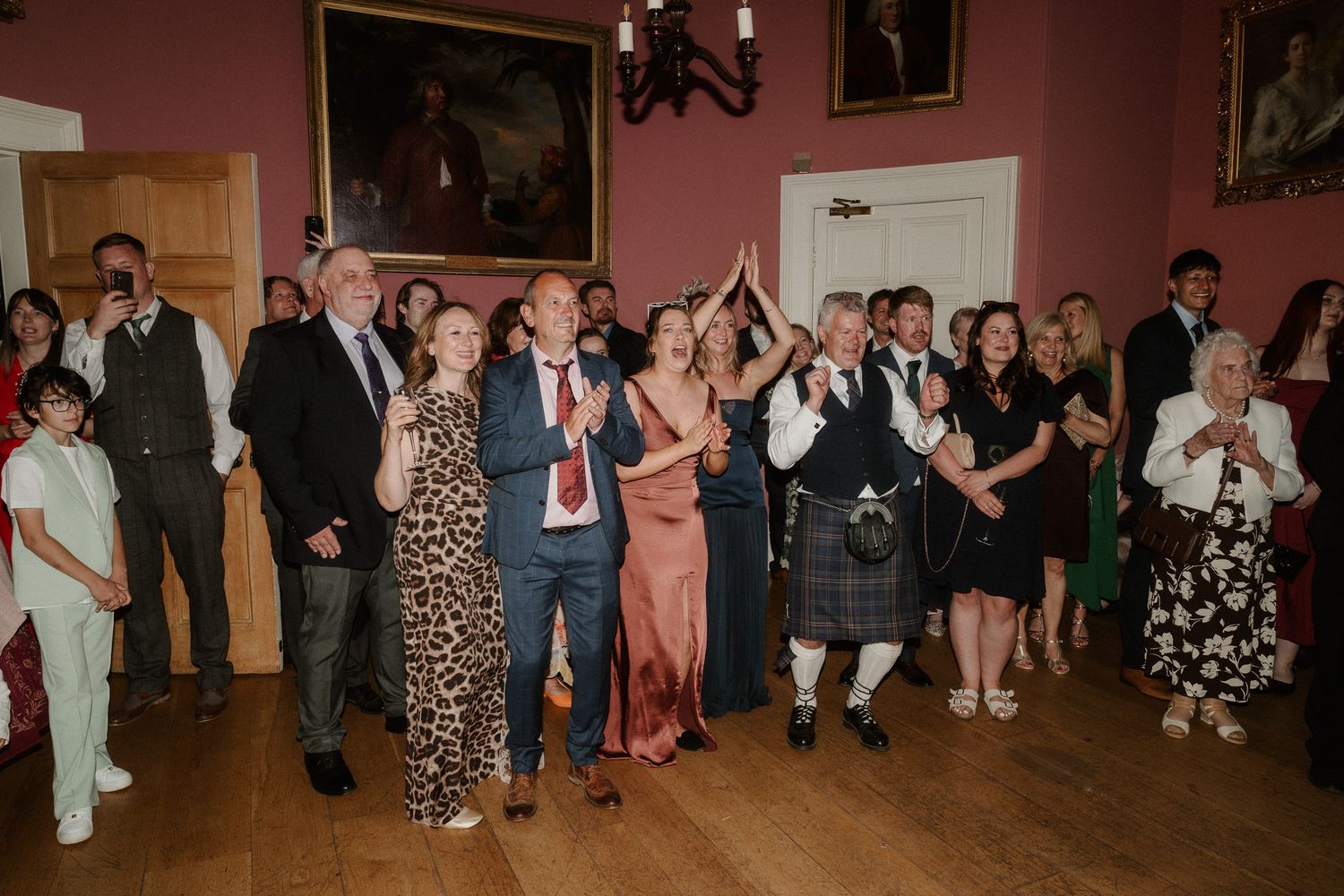 Guests clapping and dancing at a wedding reception in an elegant room, with one man wearing a traditional Scottish kilt.