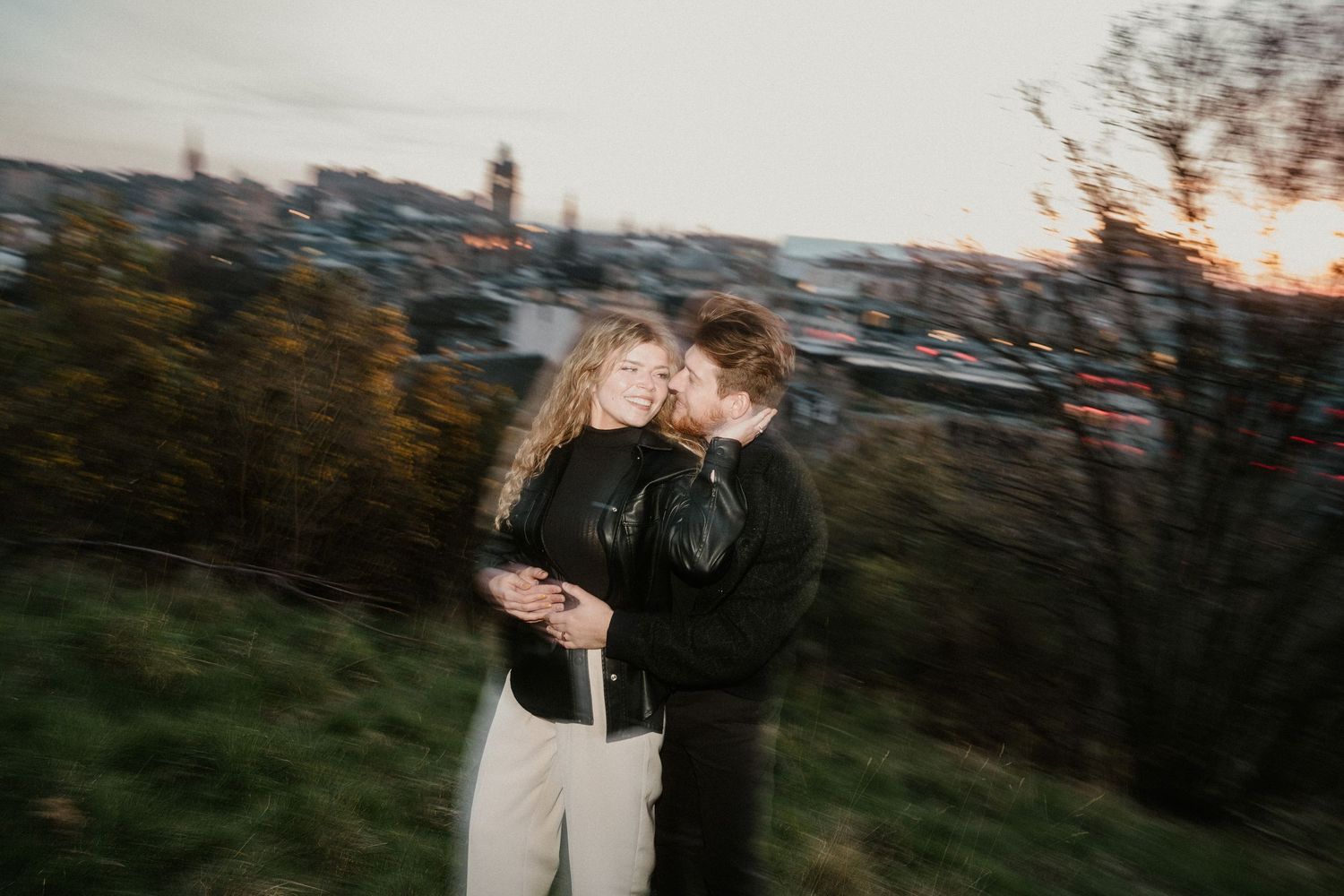 A couple embraces romantically at sunset on a hilltop overlooking a city skyline with autumn foliage.