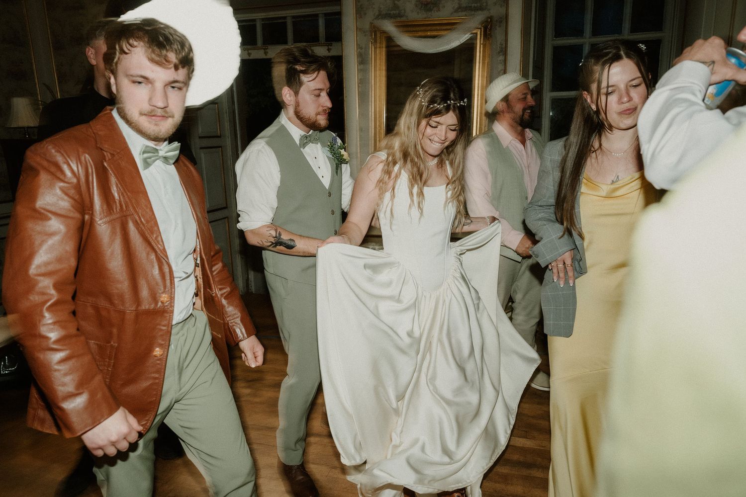 Wedding guests dancing joyfully at a reception, bride in white gown surrounded by groomsmen in sage suits.