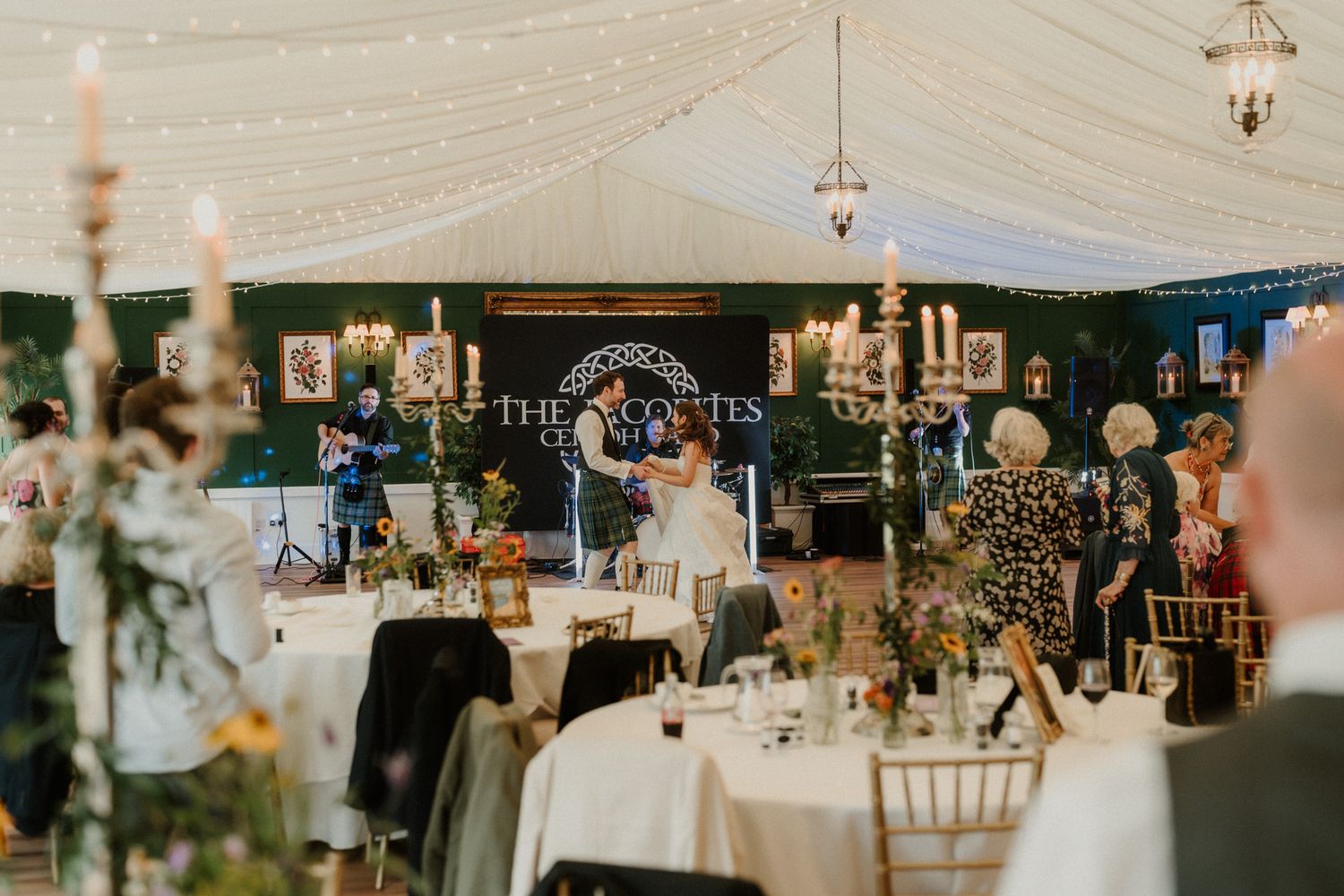 Bride and groom share first dance in a fairy-lit wedding marquee with candelabras and decorated reception tables.