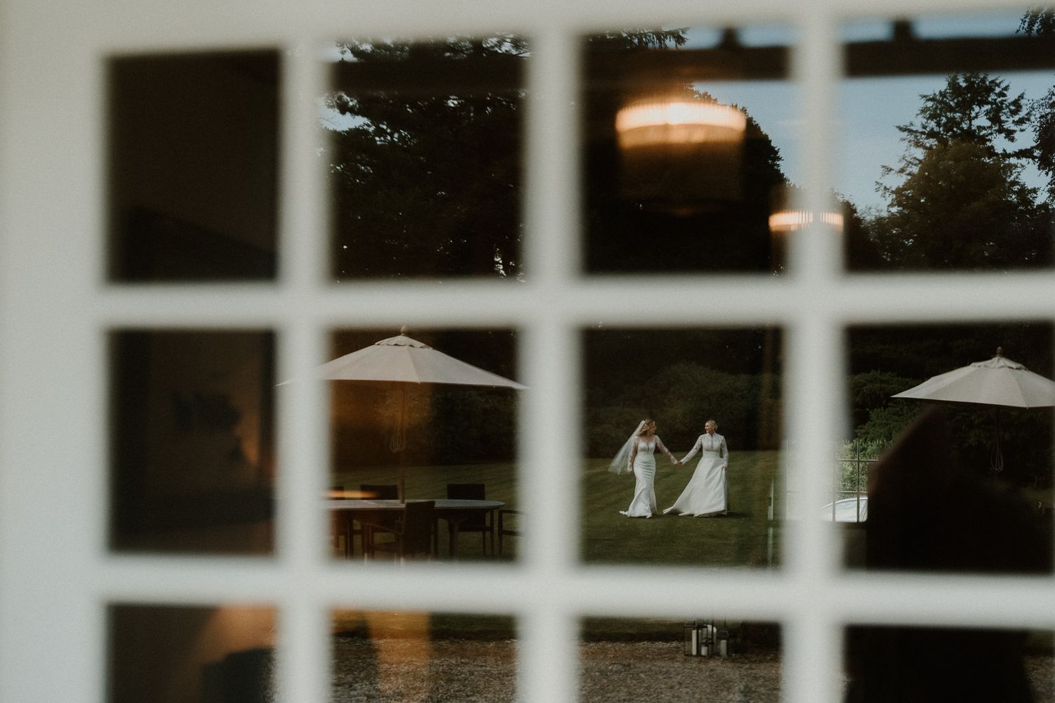 Two brides in white gowns walk across a lawn, viewed through a multi-paned window at dusk.