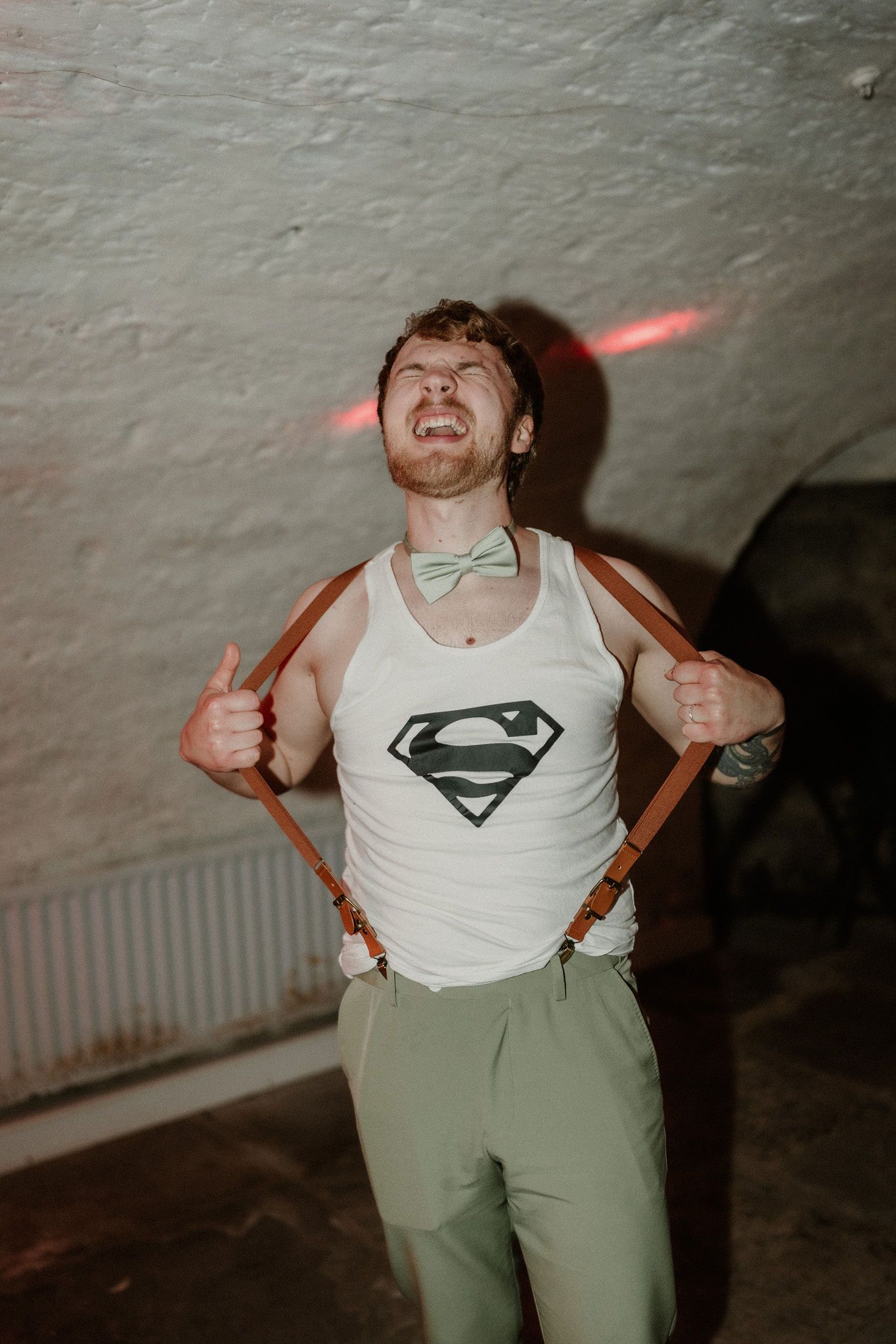 Man in Superman tank top with suspenders and bow tie giving thumbs up in dimly lit venue.