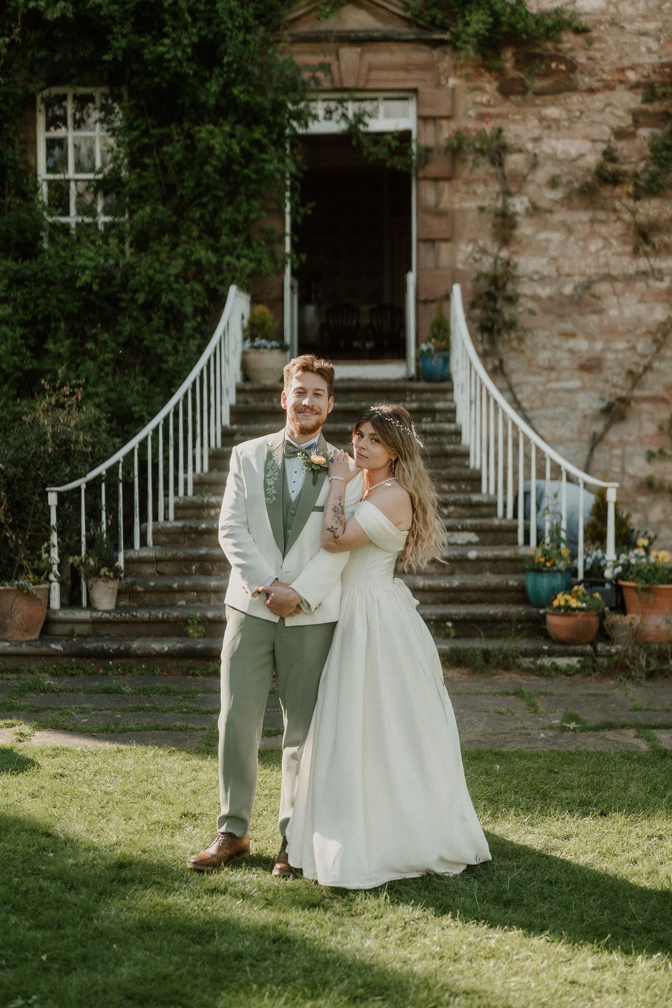 Bride and groom pose together on lawn before stone manor house steps on their wedding day.