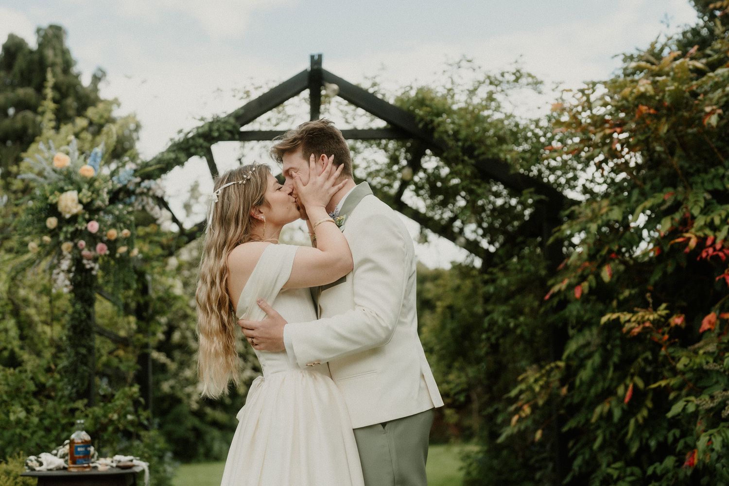 A couple shares a romantic kiss under a floral garden arch on their wedding day, surrounded by lush greenery.