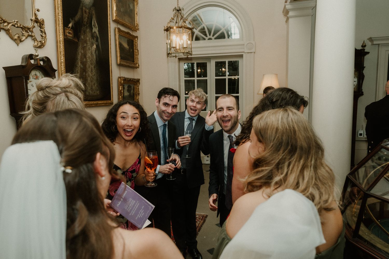 Joyful wedding guests laughing and celebrating together in an elegant venue with classic decor and warm lighting.