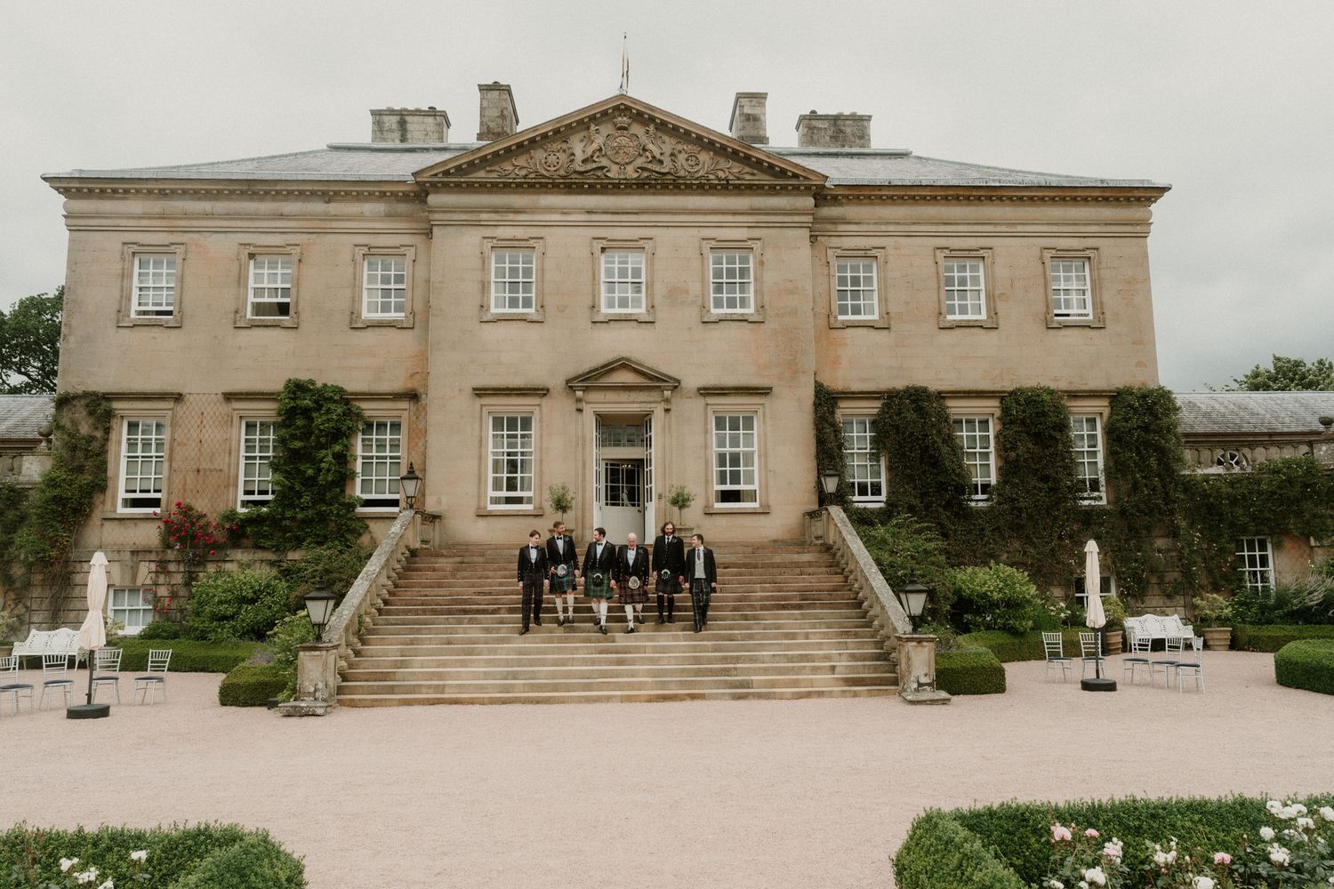 Wedding party posing on grand stone steps of elegant Georgian country house mansion surrounded by manicured gardens.