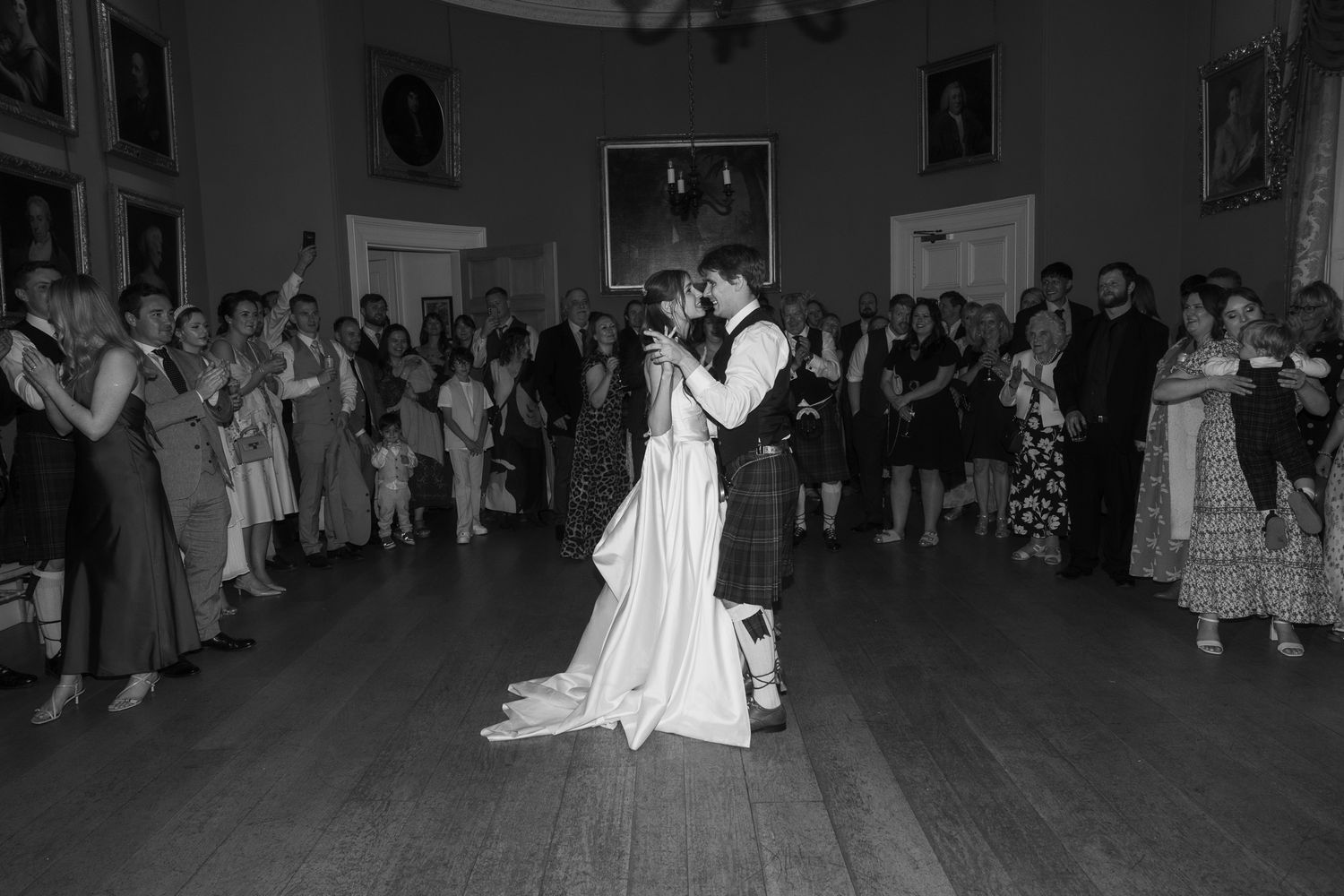 Black and white photo of a couple sharing their first dance at a wedding reception surrounded by applauding guests.