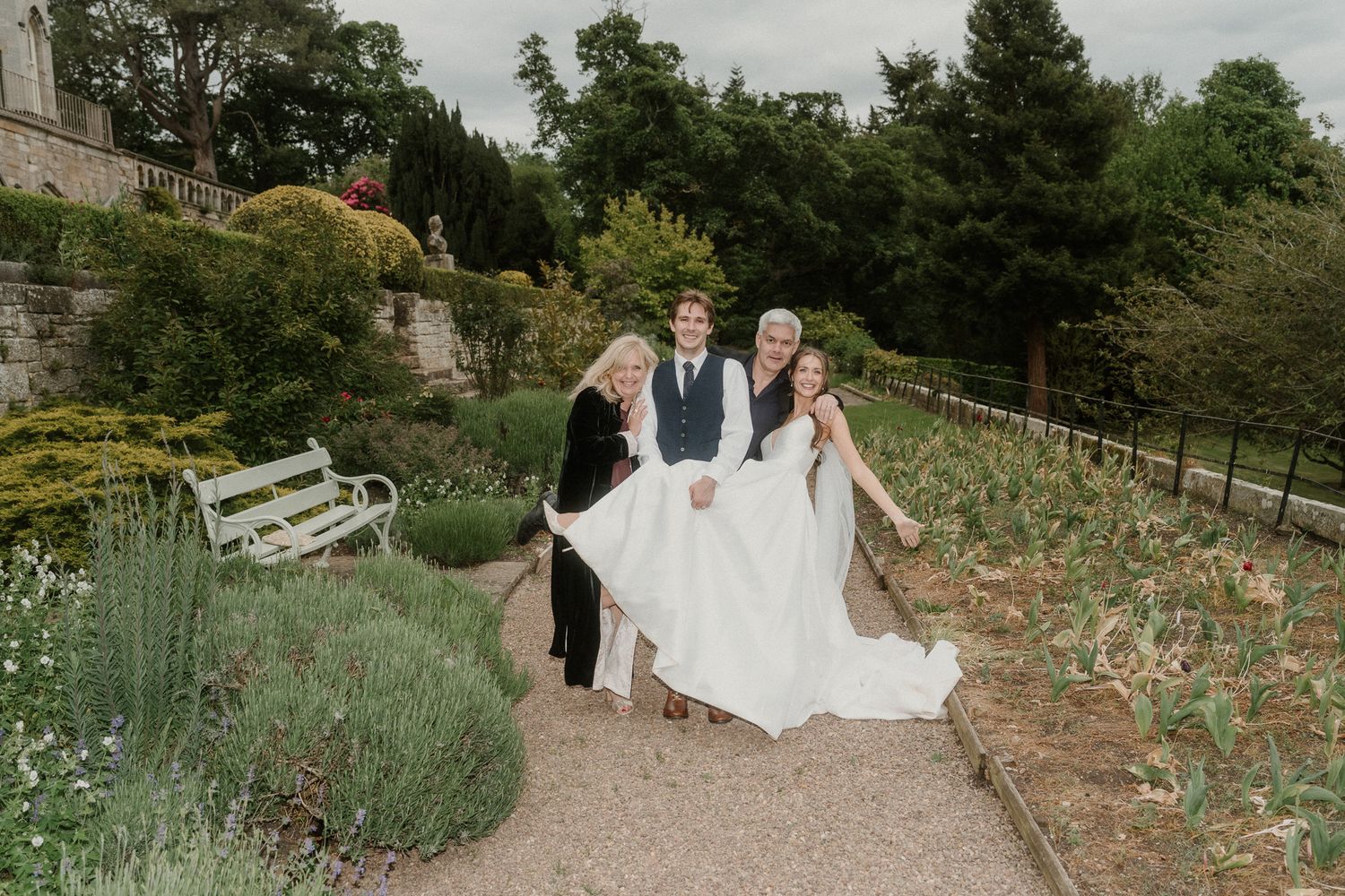 Bride in white gown poses with two guests in lush garden setting during wedding celebration outdoors.