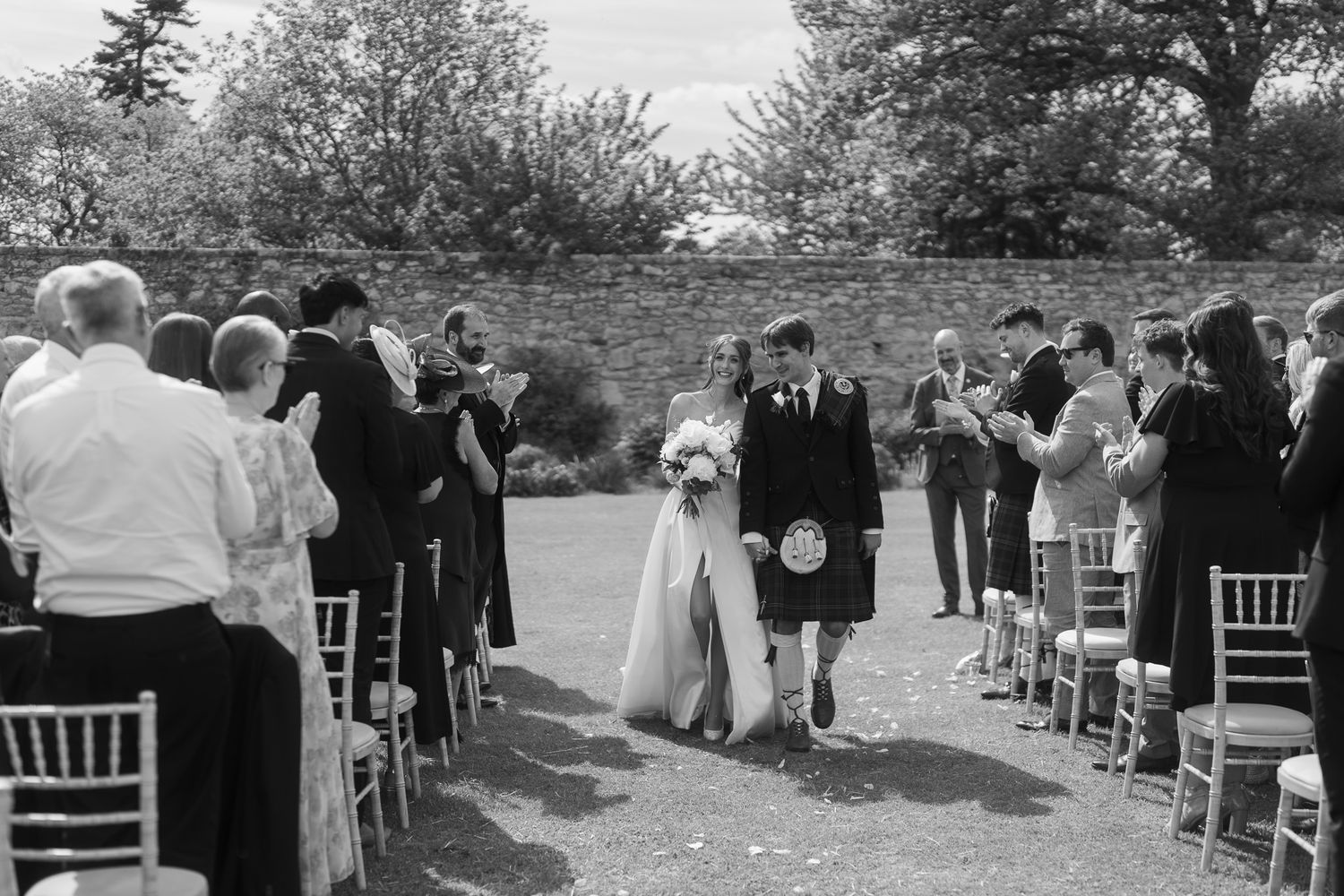 Black and white photo of bride and groom walking down outdoor wedding aisle, groom wearing Scottish kilt, guests applauding.