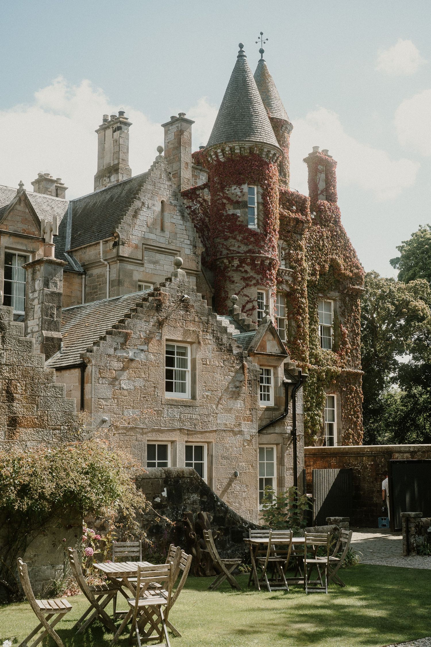 A charming Scottish castle-style stone building with turrets and ivy-covered walls surrounded by a manicured lawn.