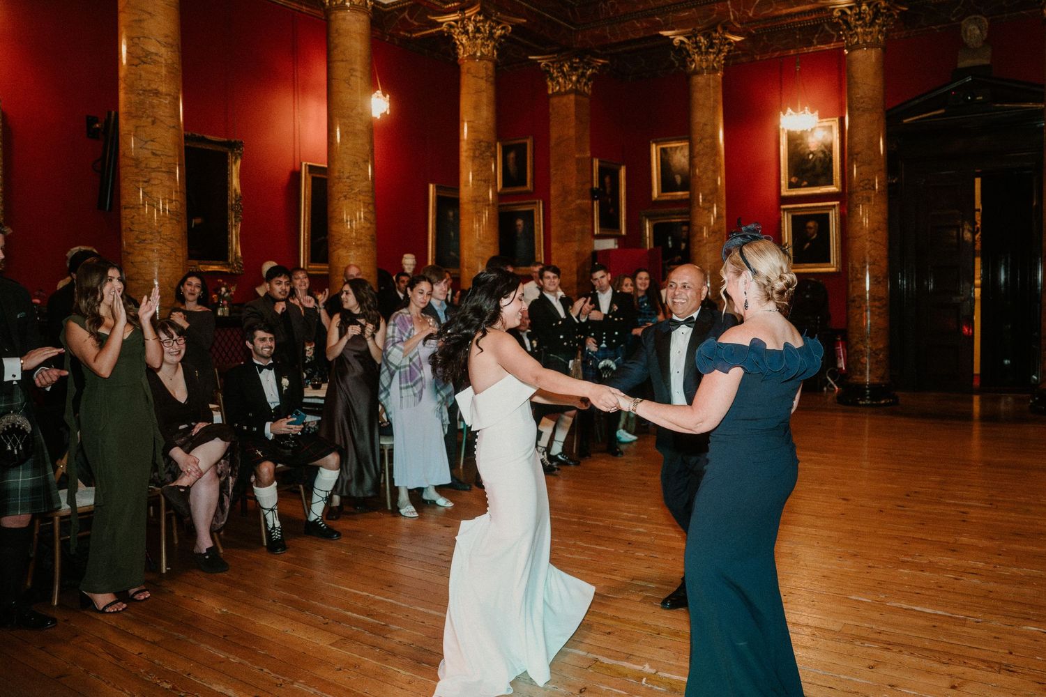 Couples dancing at a wedding reception in a grand ballroom with ornate columns and red walls.