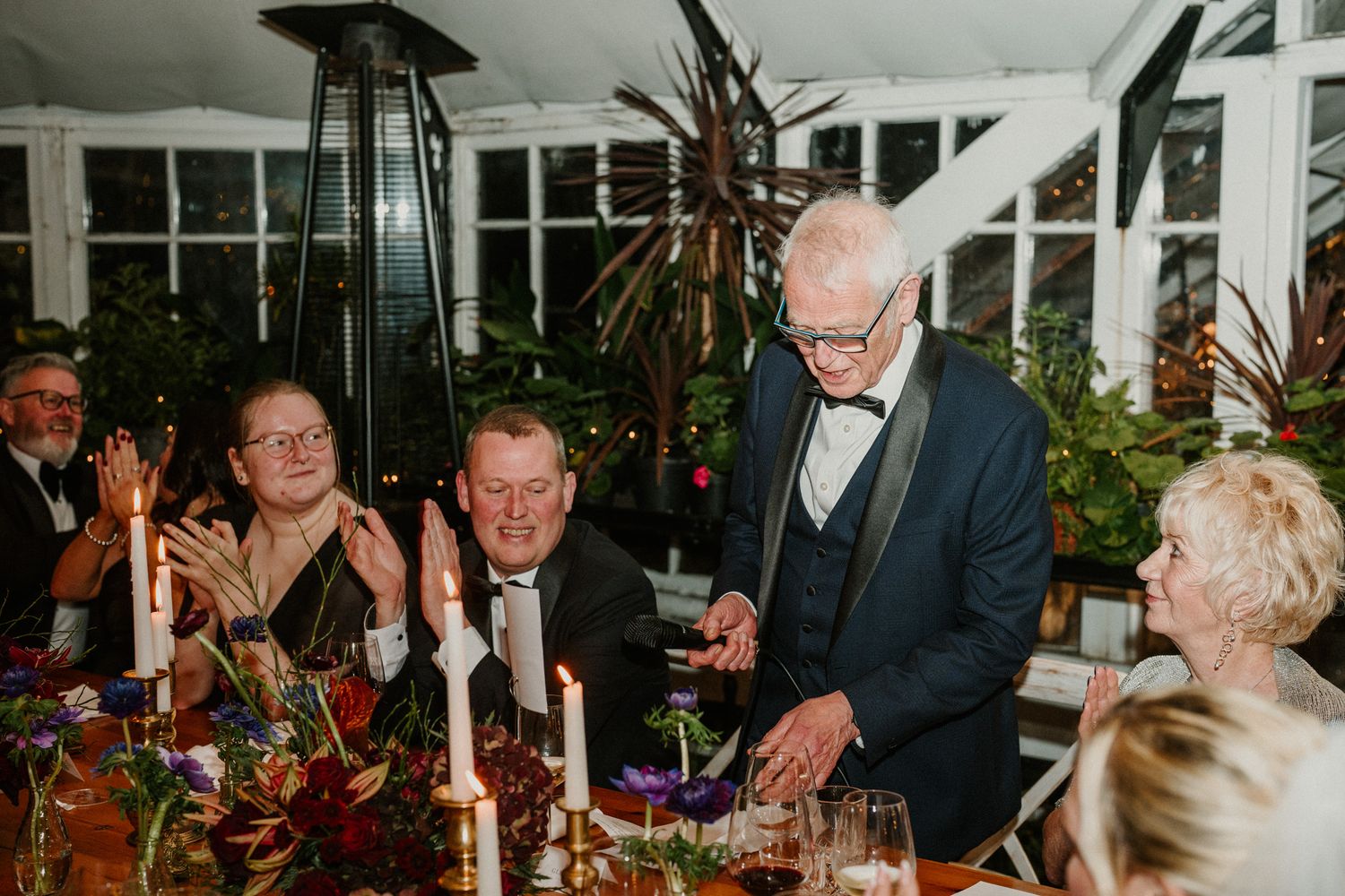 Wedding reception guests applauding during celebrations in an elegant glass-walled venue.